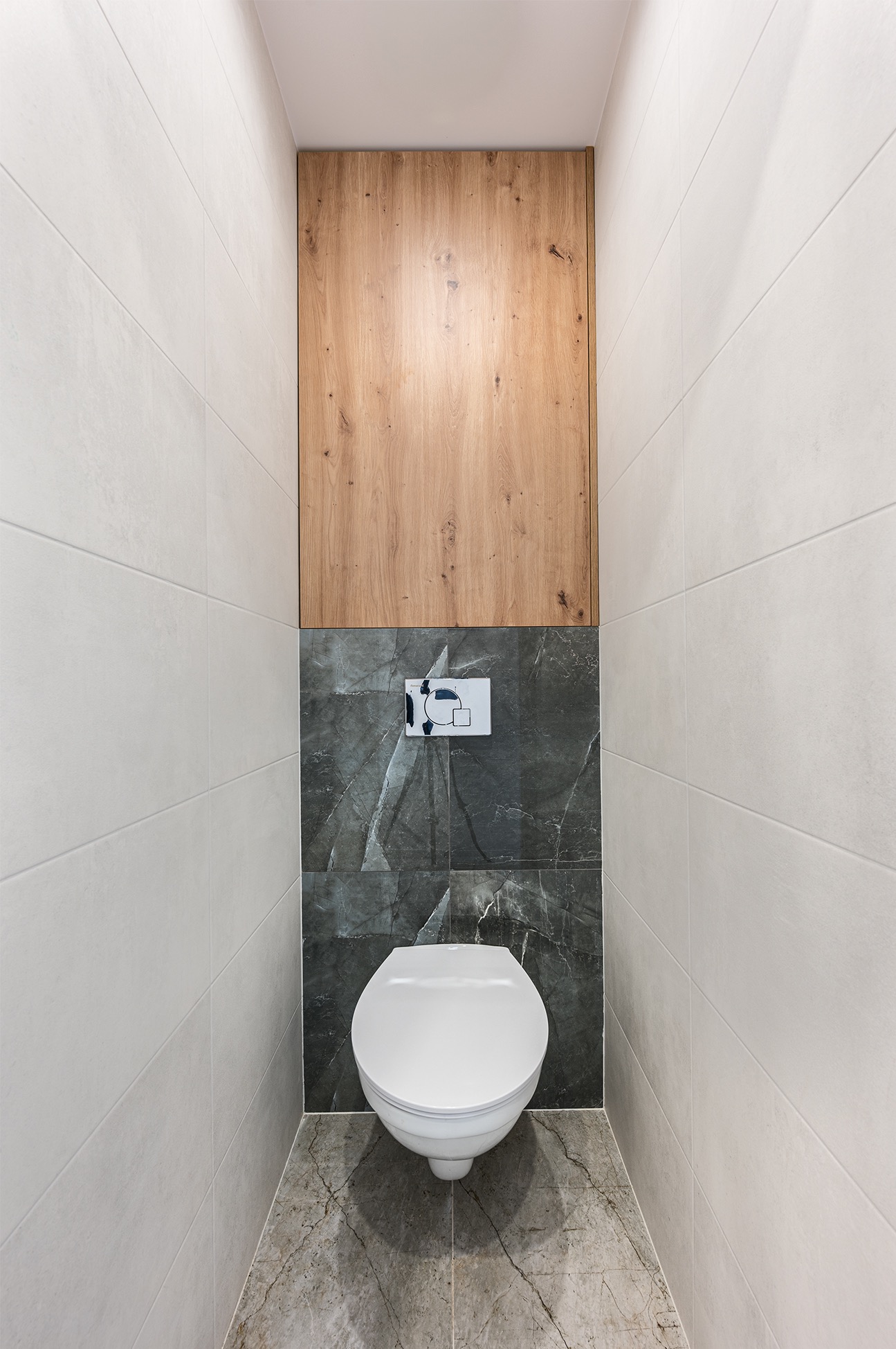 Kompaktowa toaleta w nowoczesnym stylu: biała ceramika, szare płytki imitujące marmur, drewniana zabudowa nad stelażem, minimalistyczny przycisk spłukujący.