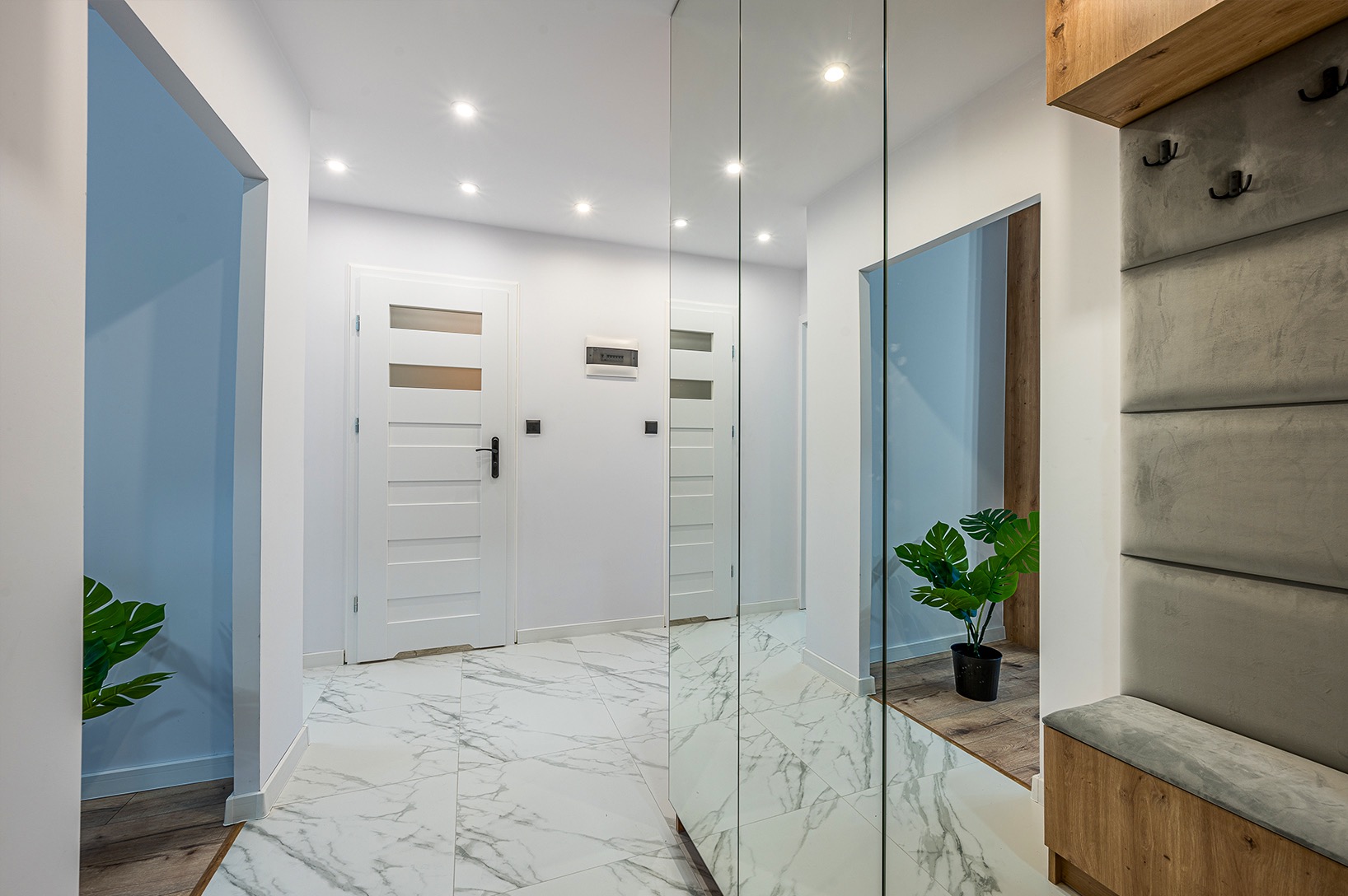 Jasne wnętrze przedpokoju z marmurową podłogą, białymi drzwiami i lustrem. Widoczna dekoracyjna roślina w doniczce i panel z wieszakami.