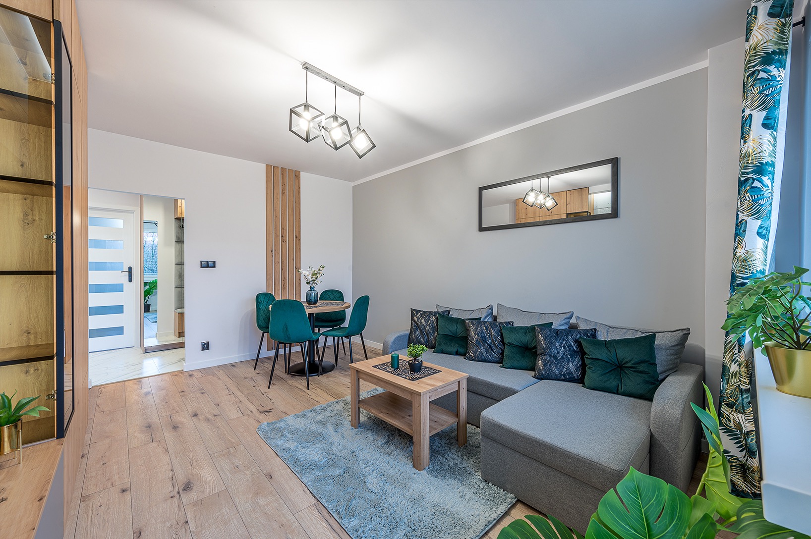 Jasne wnętrze salonu z szarą sofą narożną, drewnianym stolikiem kawowym na tle turkusowego dywanu i okrągłym stołem z zielonymi krzesłami. Widoczna drewniana podłoga i dekoracje.