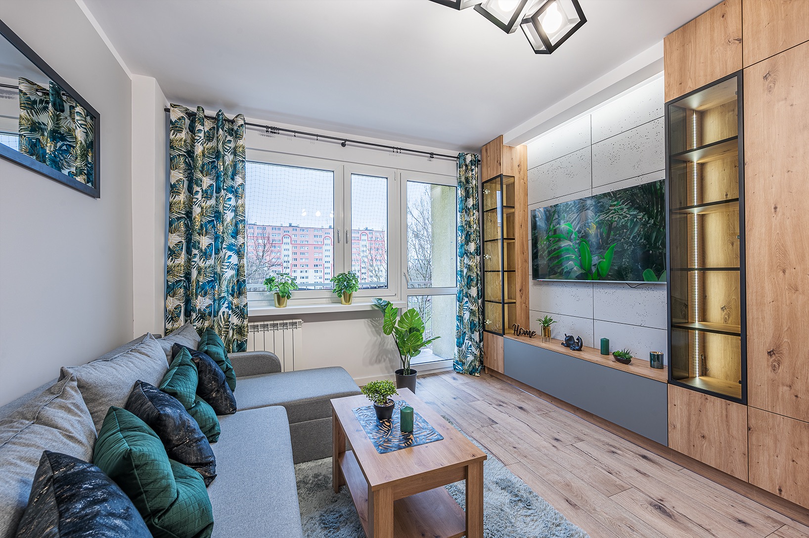 Jasne wnętrze salonu z szarą sofą, drewnianym stolikiem kawowym i ścianą z telewizorem otoczoną drewnianą zabudową. Okno z roślinami i wzorzyste zasłony dodają koloru.