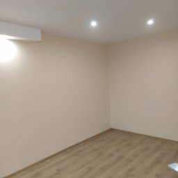 Green Work - Jasne wnętrze po malowaniu: beżowe ściany, drewnopodobna podłoga, sufit z oświetleniem punktowym i klimatyzacją. Widok na róg pokoju.