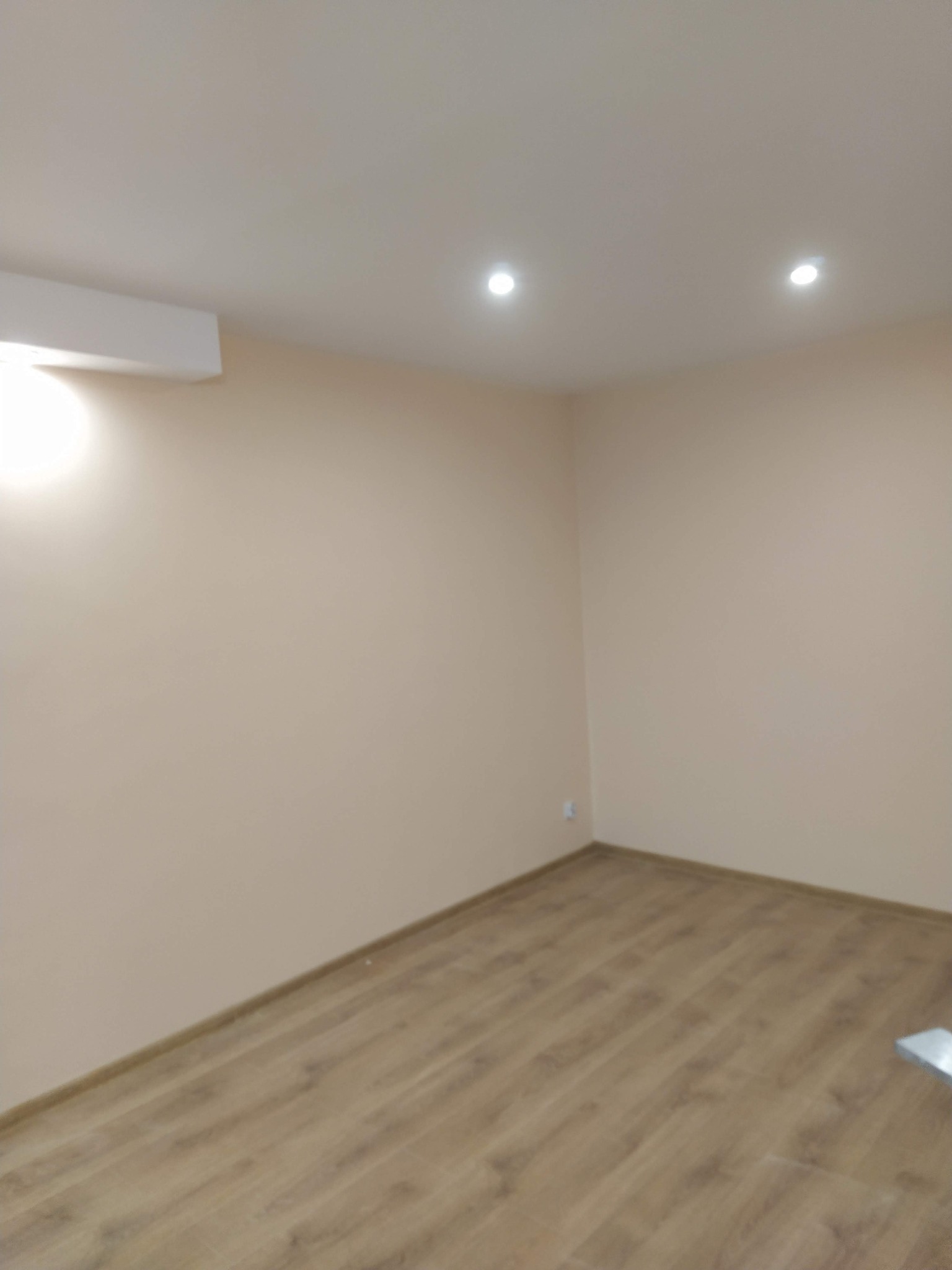 Jasne wnętrze po malowaniu: beżowe ściany, drewnopodobna podłoga, sufit z oświetleniem punktowym i klimatyzacją. Widok na róg pokoju.