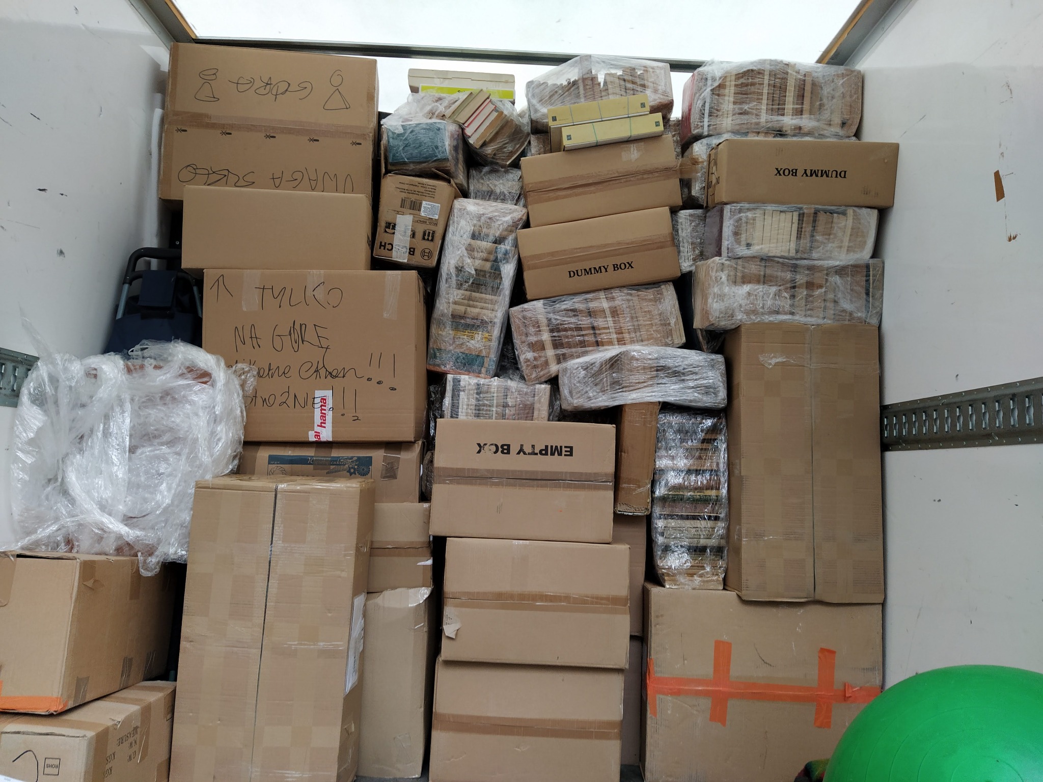 Wnętrze furgonetki wypełnionej kartonami różnej wielkości, niektóre z napisami i rysunkami, inne owinięte folią, sugerujące transport lub przeprowadzkę.