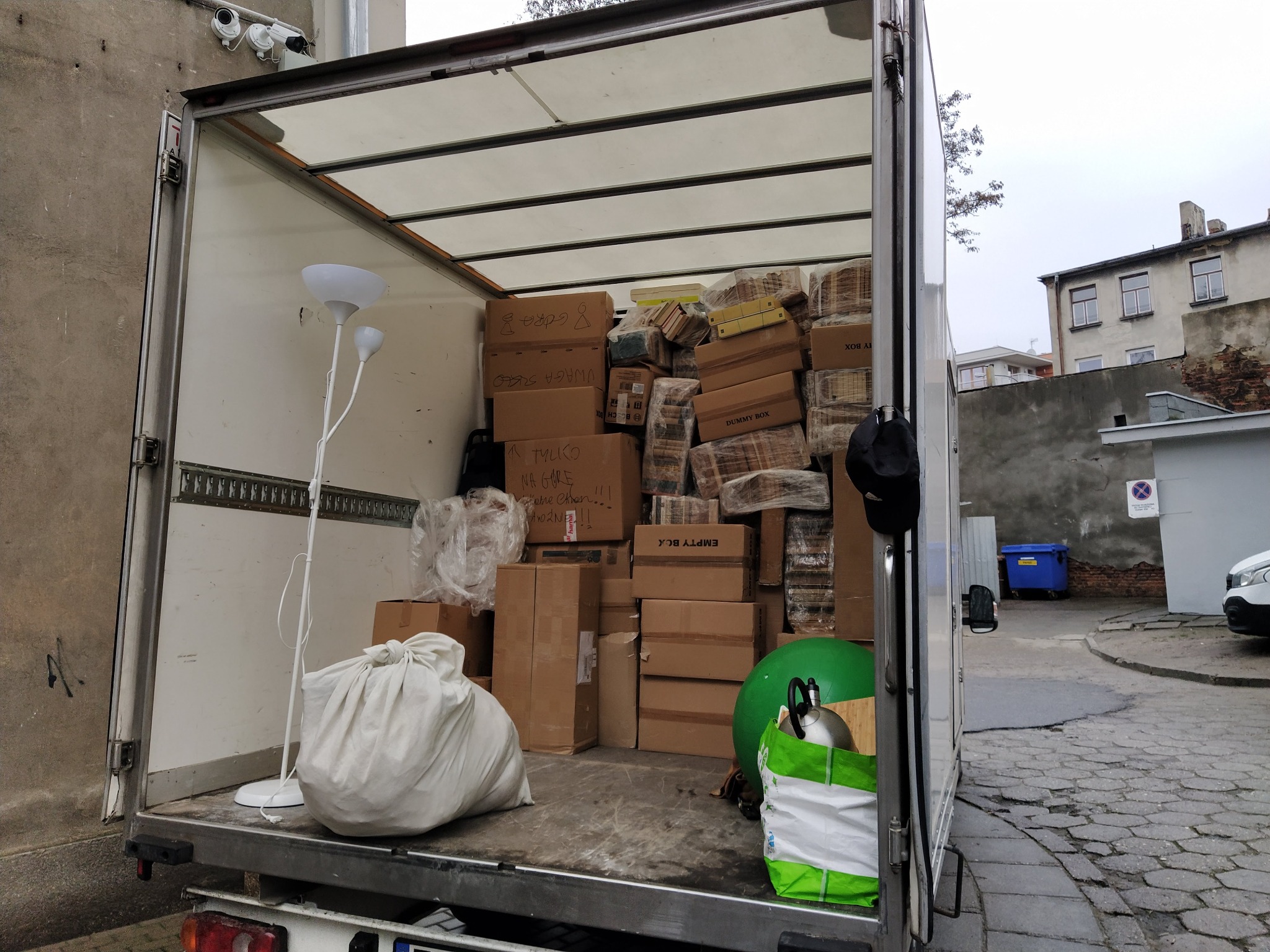 Wnętrze furgonetki wypełnionej kartonami, workami i lampą podłogową, gotowe do transportu. Widoczne budynki w tle.