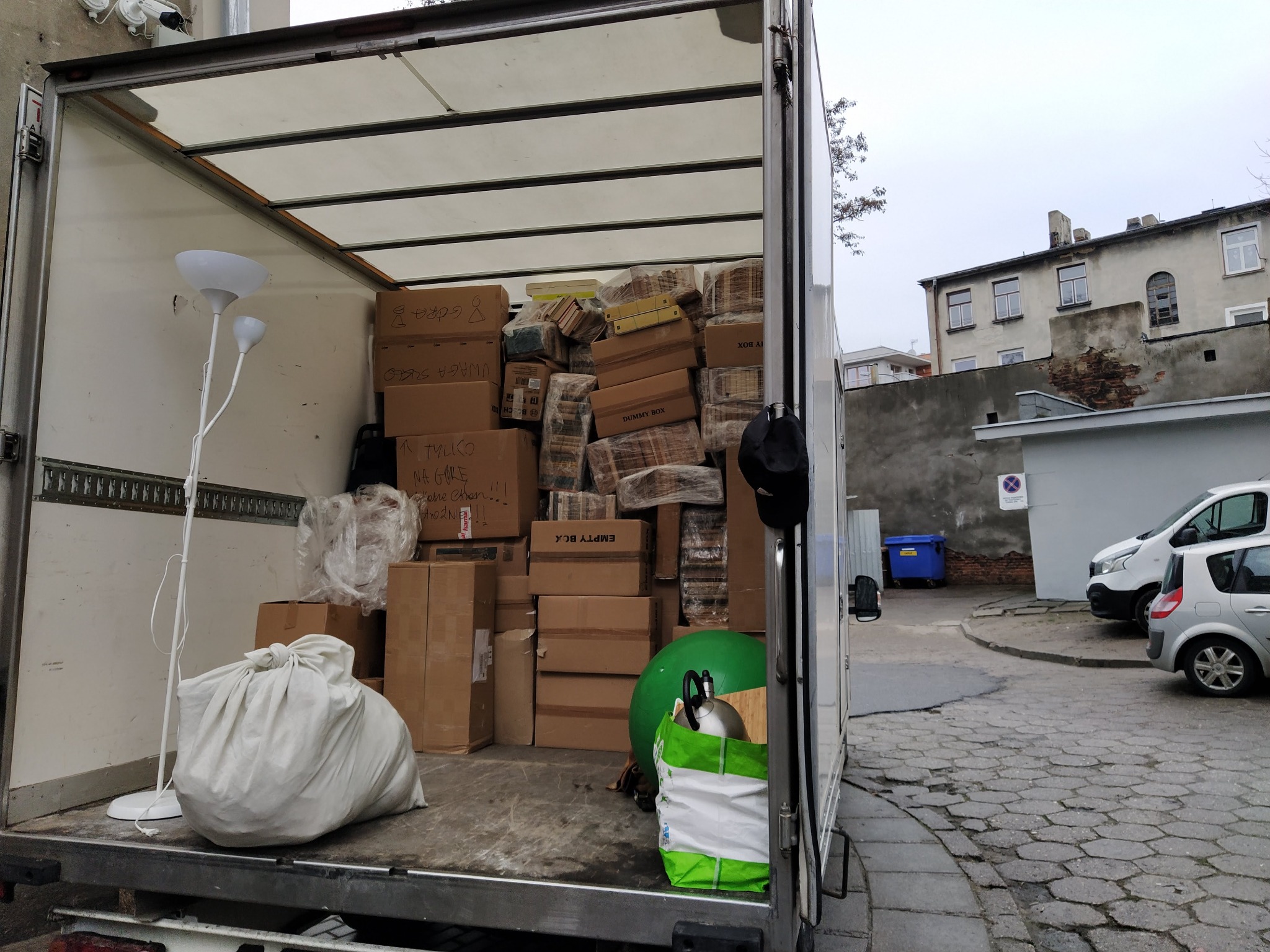 Wnętrze furgonetki wypełnionej kartonami, lampą i workiem, gotowe do transportu. Widoczne budynki w tle.