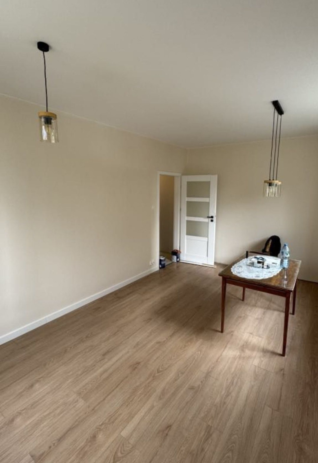 Jasne, puste wnętrze pokoju po malowaniu ścian i położeniu paneli podłogowych. Widoczne lampy, drzwi i stół z przedmiotami. Świeżo wyremontowane pomieszczenie.