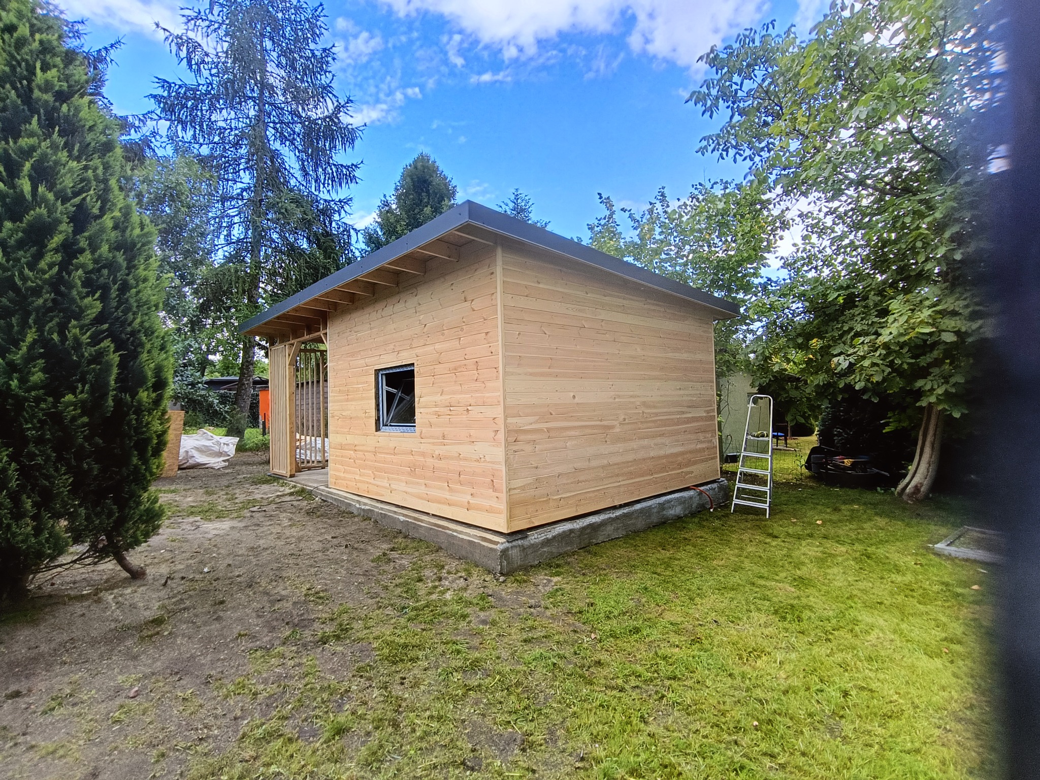 Nowo wybudowany, drewniany domek narzędziowy z czarną ramą okienną, na betonowej podstawie, otoczony zielenią i drzewami, z drabiną opartą o ścianę.