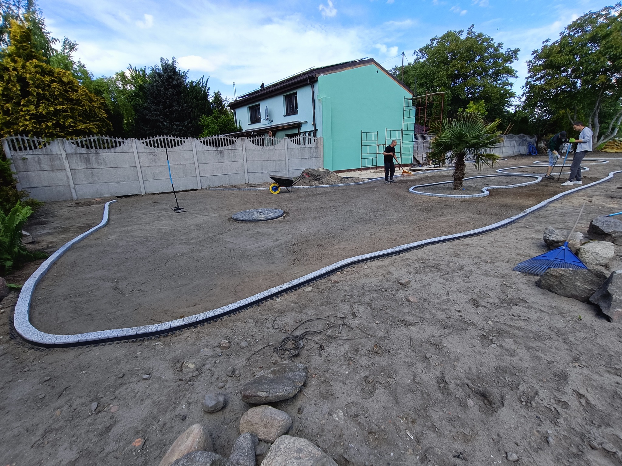 Prace ziemne w ogrodzie: układanie obrzeży z kostki, wyrównywanie terenu pod przyszły trawnik. W tle dom z rusztowaniem i pracownicy. Widoczne narzędzia ogrodnicze.