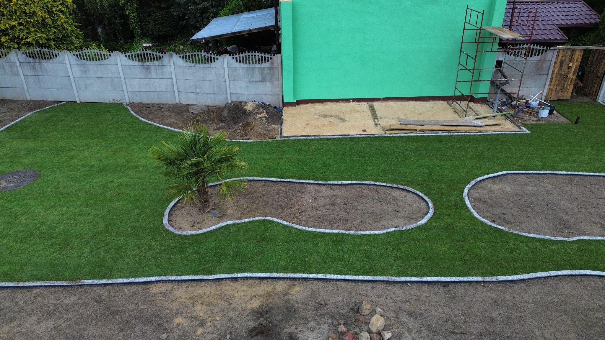 Widok z góry na świeżo założony trawnik z palmą i rabatami obrzeżonymi kamieniem, przylegający do budynku z zieloną elewacją i betonowym ogrodzeniem.