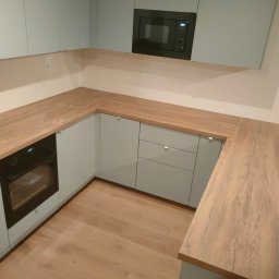 Eniu Design Ernest Borowiak - Jasna kuchnia w kształcie litery L z drewnianym blatem, wbudowanym piekarnikiem i mikrofalówką. Minimalistyczny design, drewniana podłoga, neutralne kolory.