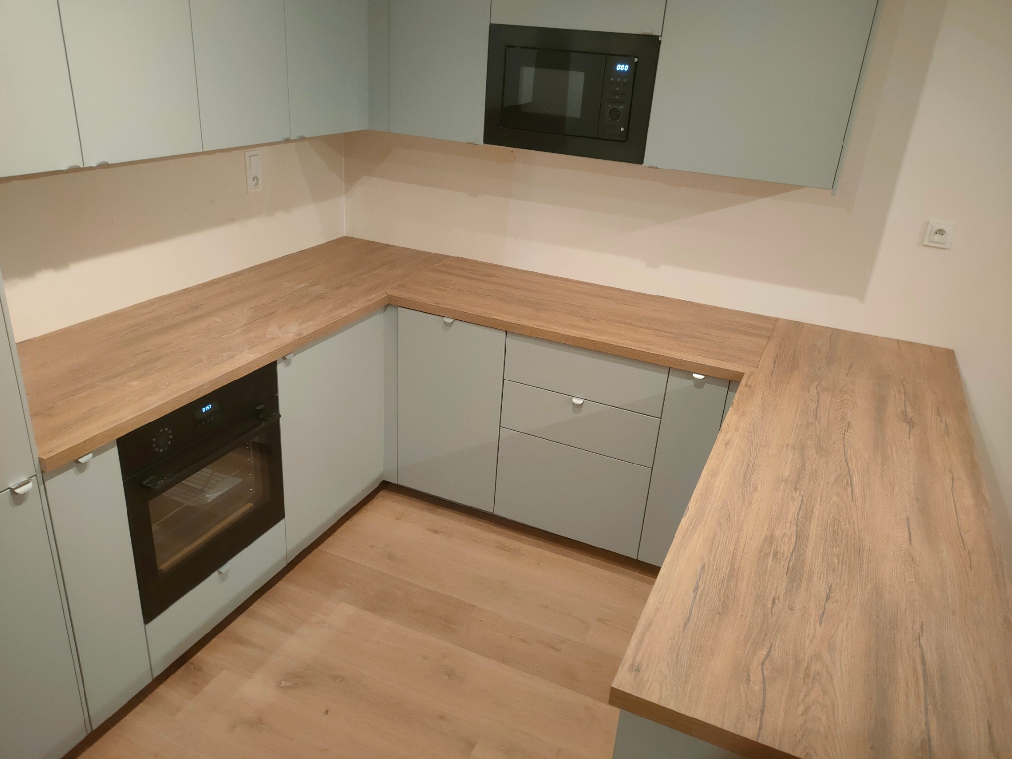 Jasna kuchnia w kształcie litery L z drewnianym blatem, wbudowanym piekarnikiem i mikrofalówką. Minimalistyczny design, drewniana podłoga, neutralne kolory.