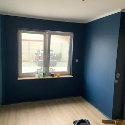 AK-BUD - Pomieszczenie po malowaniu ścian na granatowo, widoczne narzędzia malarskie i podłoga z paneli. Przez okno widać samochód i betonowy płot.