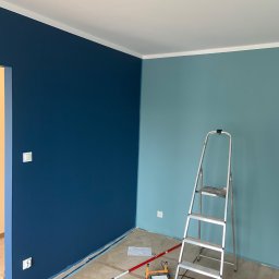 AK-BUD - Ściana pomalowana na dwa kolory: granatowy i błękitny. Widoczna drabina malarska, wałek i inne narzędzia malarskie. Podłoga zabezpieczona folią malarską.