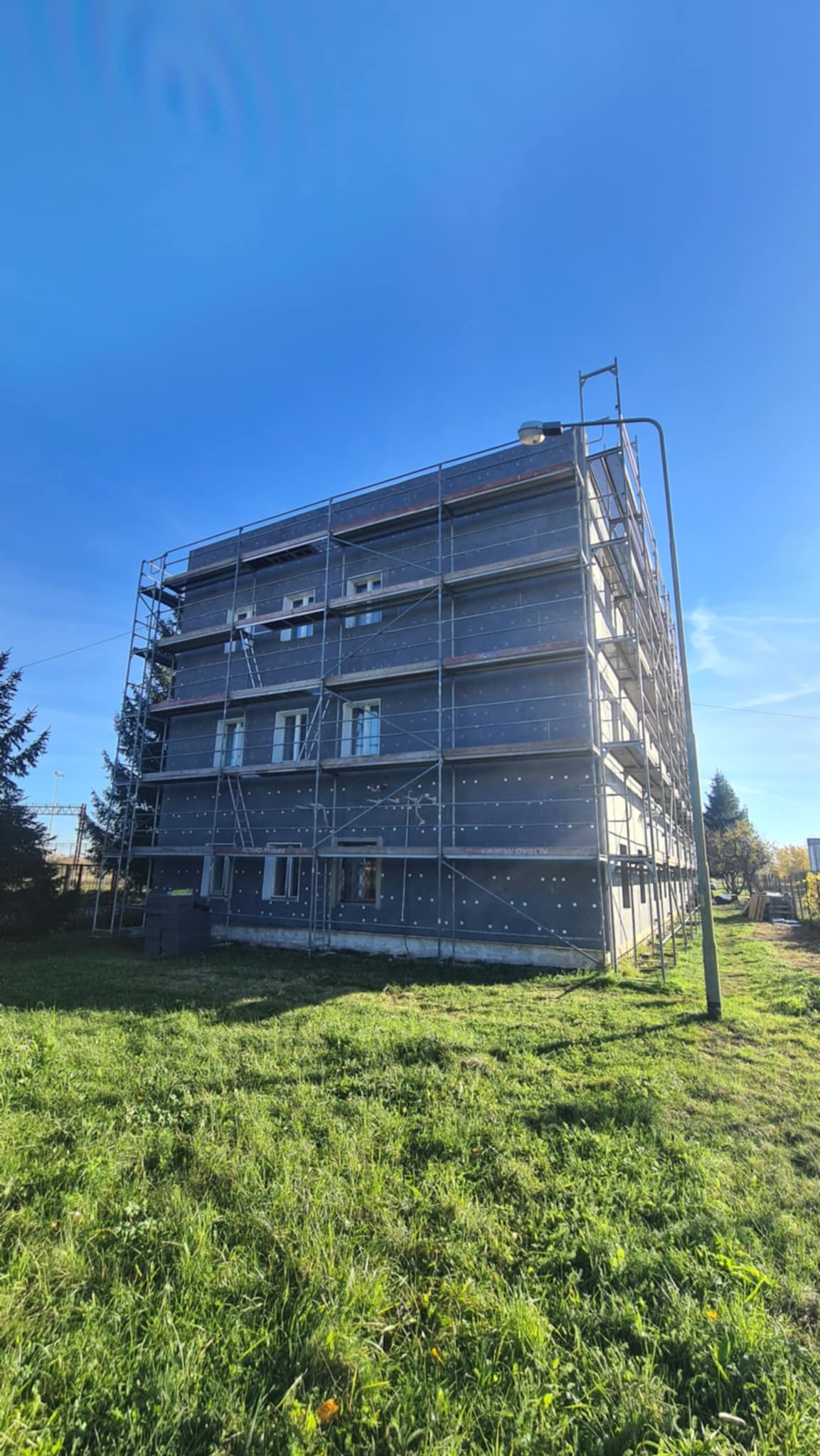 Budynek wielorodzinny w trakcie ocieplania, osłonięty rusztowaniami. Widoczne płyty izolacyjne na elewacji, błękitne niebo i trawnik. Ujęcie z dołu.