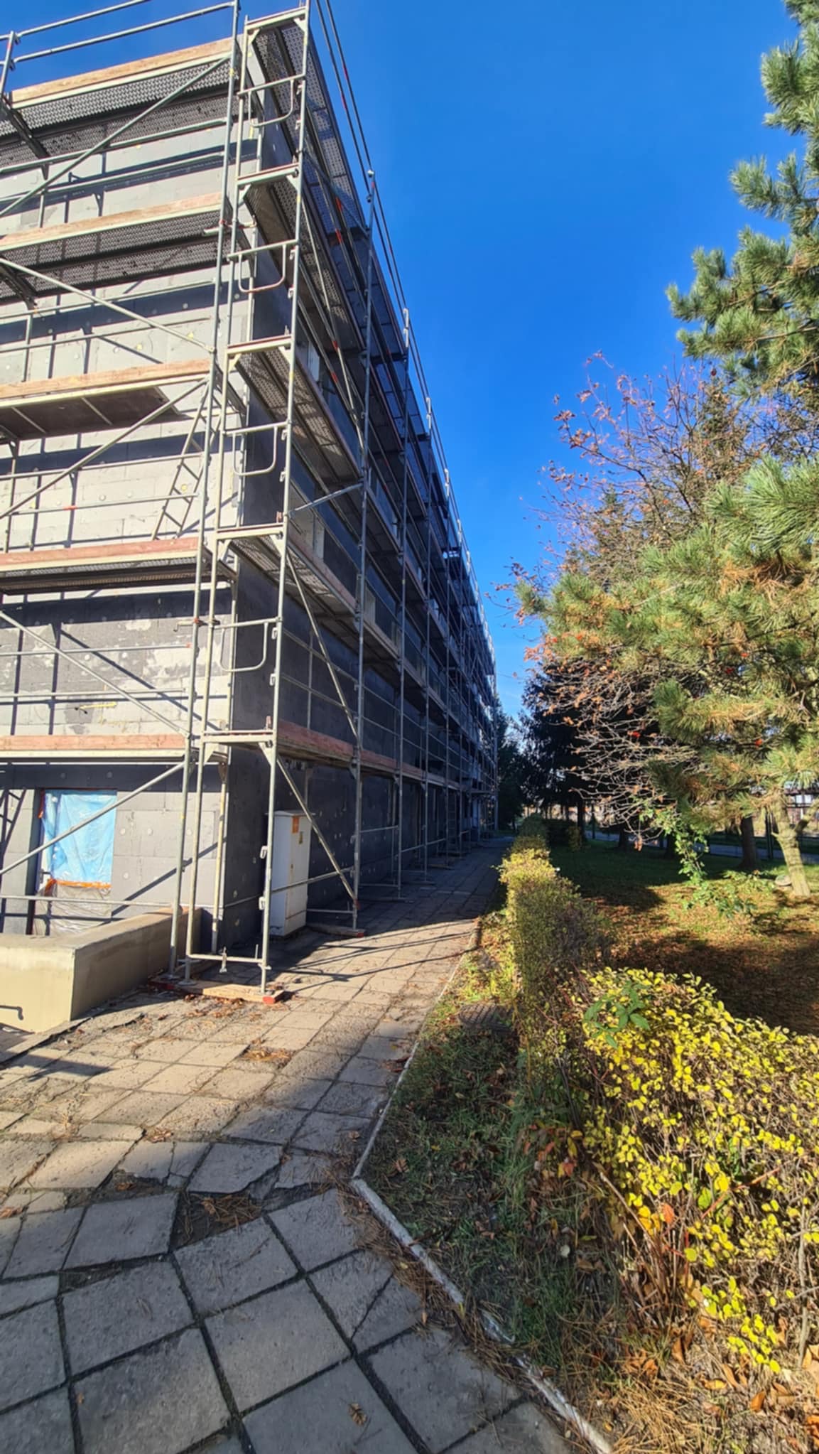 Budynek w trakcie prac elewacyjnych, osłonięty rusztowaniami na tle błękitnego nieba i zieleni drzew. Widoczny fragment chodnika i krzewów.