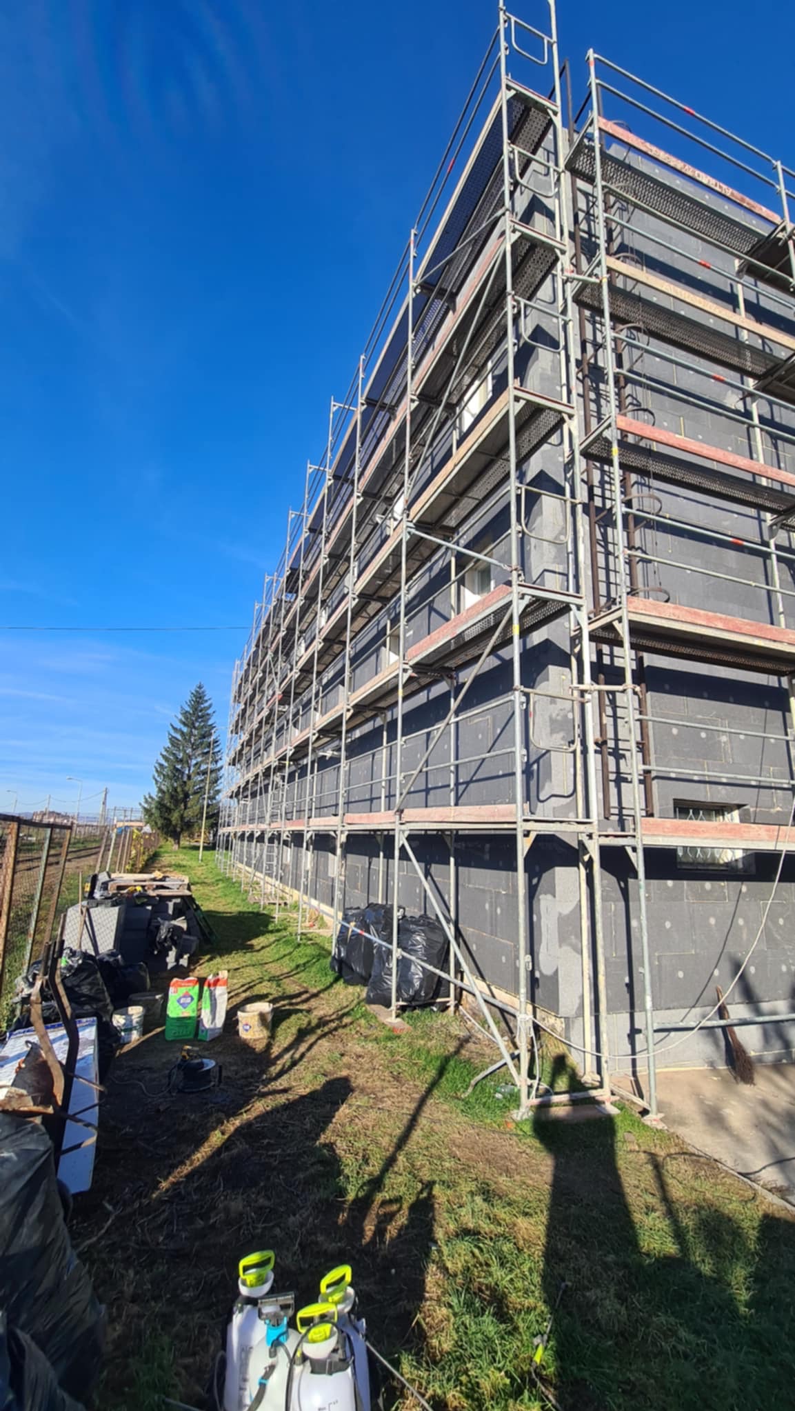 Budynek wielorodzinny w trakcie ocieplania, osłonięty rusztowaniami. Widoczne materiały budowlane i sprzęt. Słoneczny dzień, błękitne niebo.