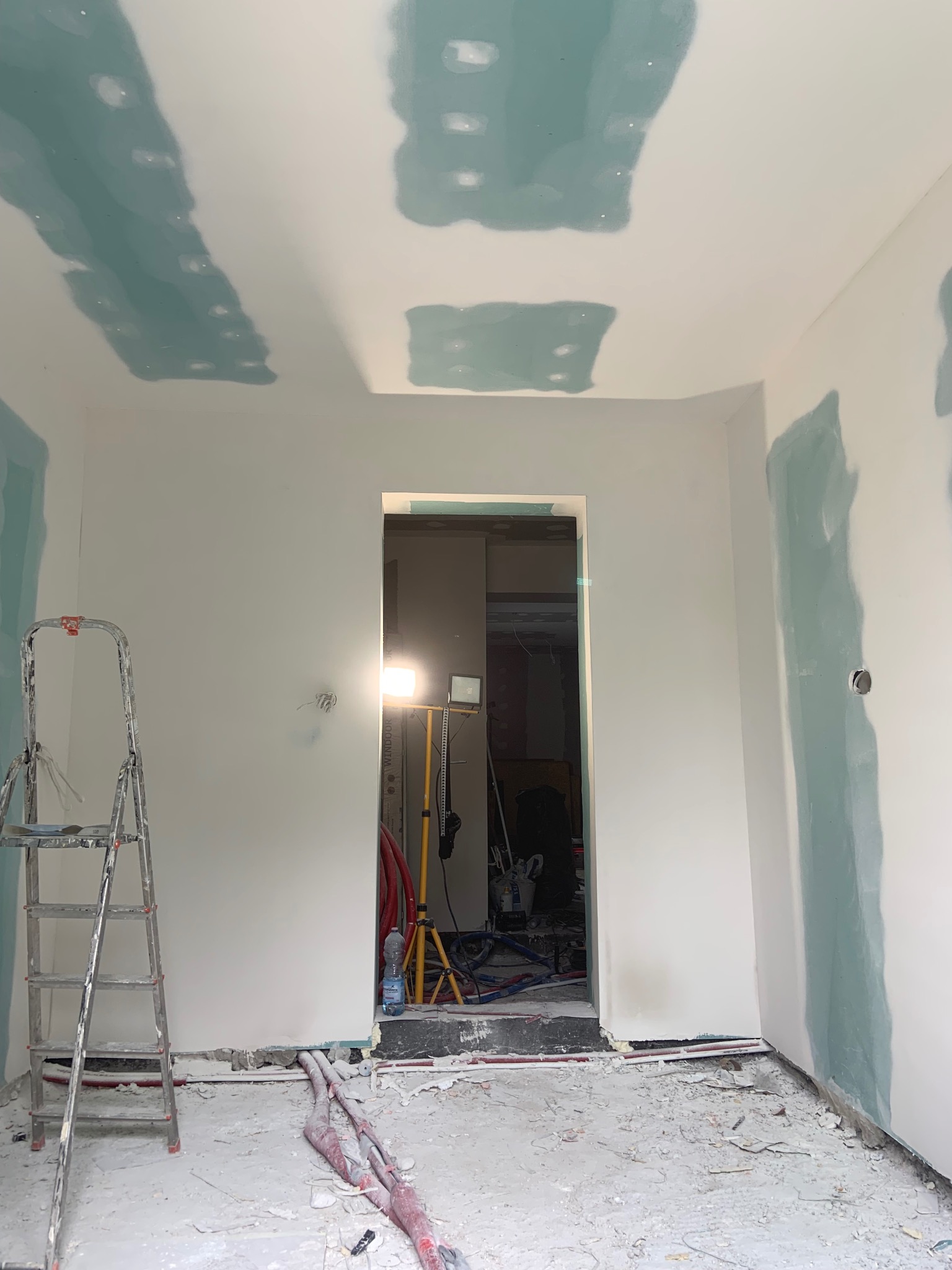Pokój w trakcie prac przygotowawczych: ściany i sufit pokryte szpachlą, widoczna drabina, instalacje i oświetlenie. Przejście do kolejnego pomieszczenia w tle.
