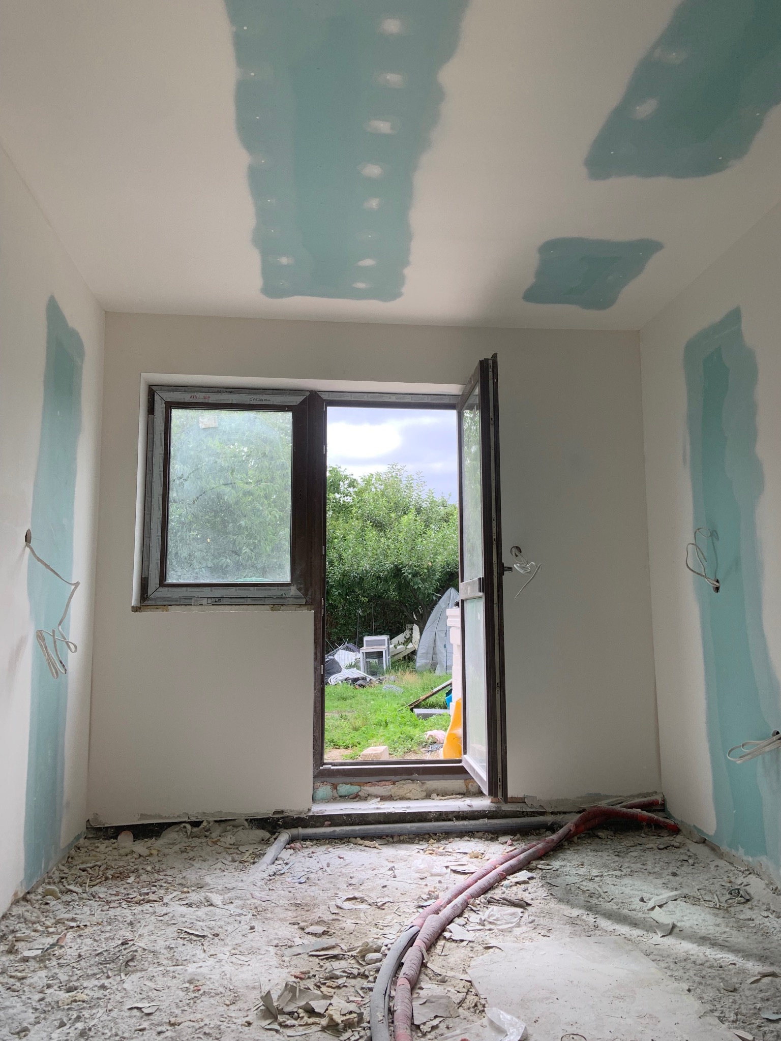 Pokój w trakcie remontu: ściany z fragmentami szpachli, otwarte okno z widokiem na zieleń, podłoga pokryta gruzem i wystające kable. Widoczne ślady po instalacji elektrycznej.