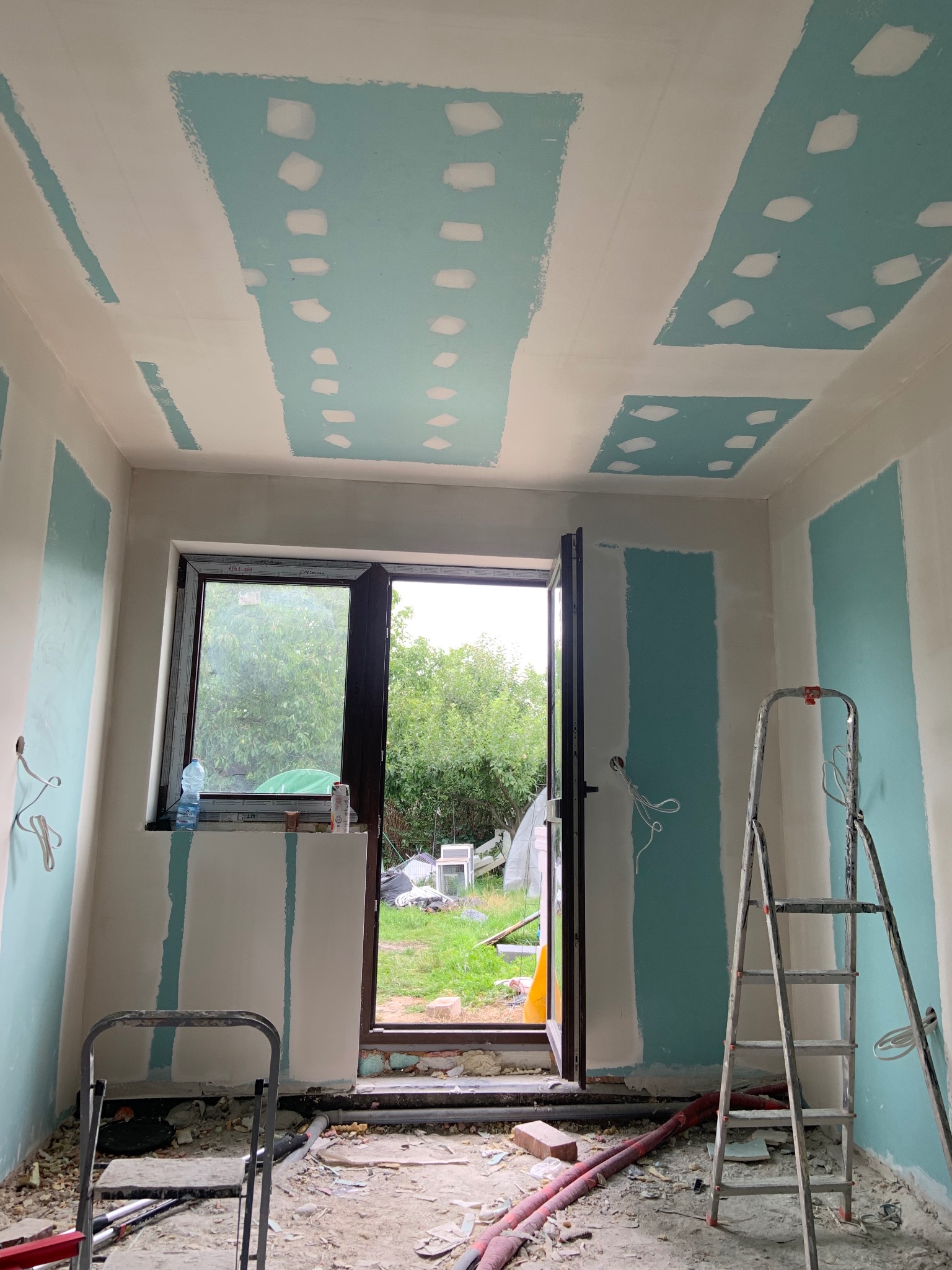 Wnętrze w trakcie prac przygotowawczych: ściany z fragmentami niebieskiej farby, sufit z zaznaczonymi miejscami na płyty, drabina i otwarte drzwi na ogród.