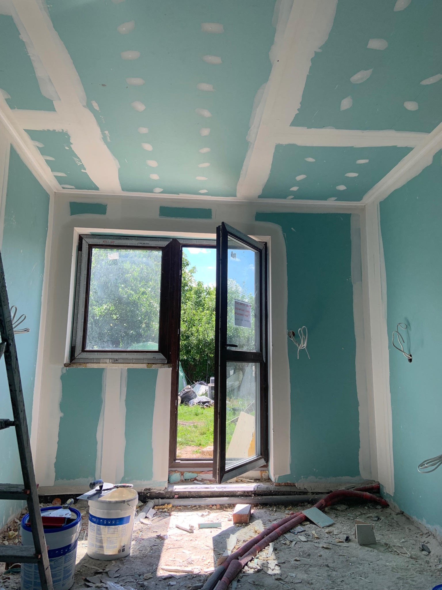 Pokój w trakcie prac wykończeniowych: ściany z płyt gipsowo-kartonowych, częściowo wyszpachlowane, otwarte okno z widokiem na zewnątrz, widoczne narzędzia i materiały budowlane.