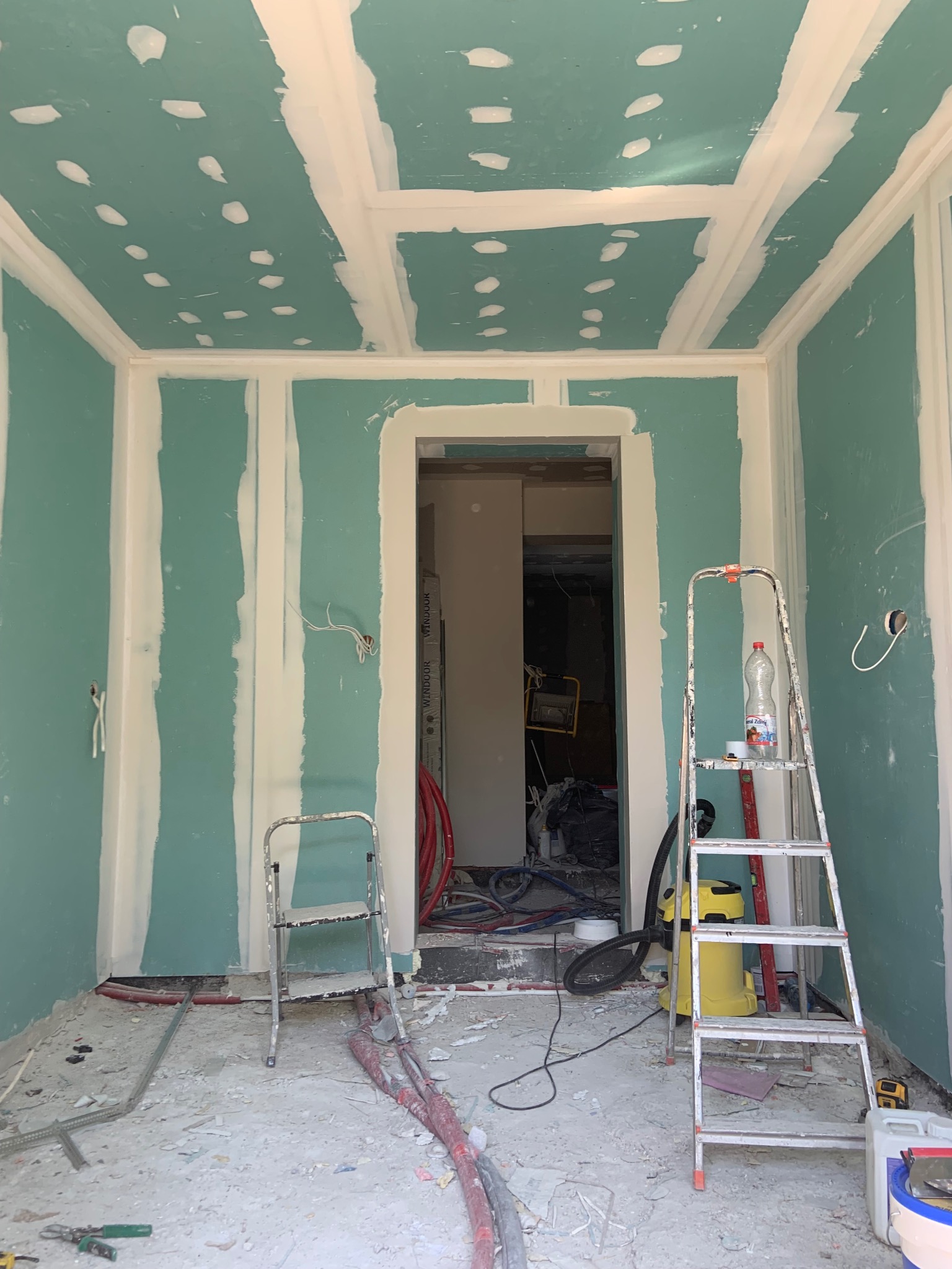 Pokój w trakcie prac wykończeniowych: płyty gipsowo-kartonowe na ścianach i suficie, szpachlowane łączenia, drabina, widoczne instalacje. W tle otwarte przejście do innego pomieszczenia.