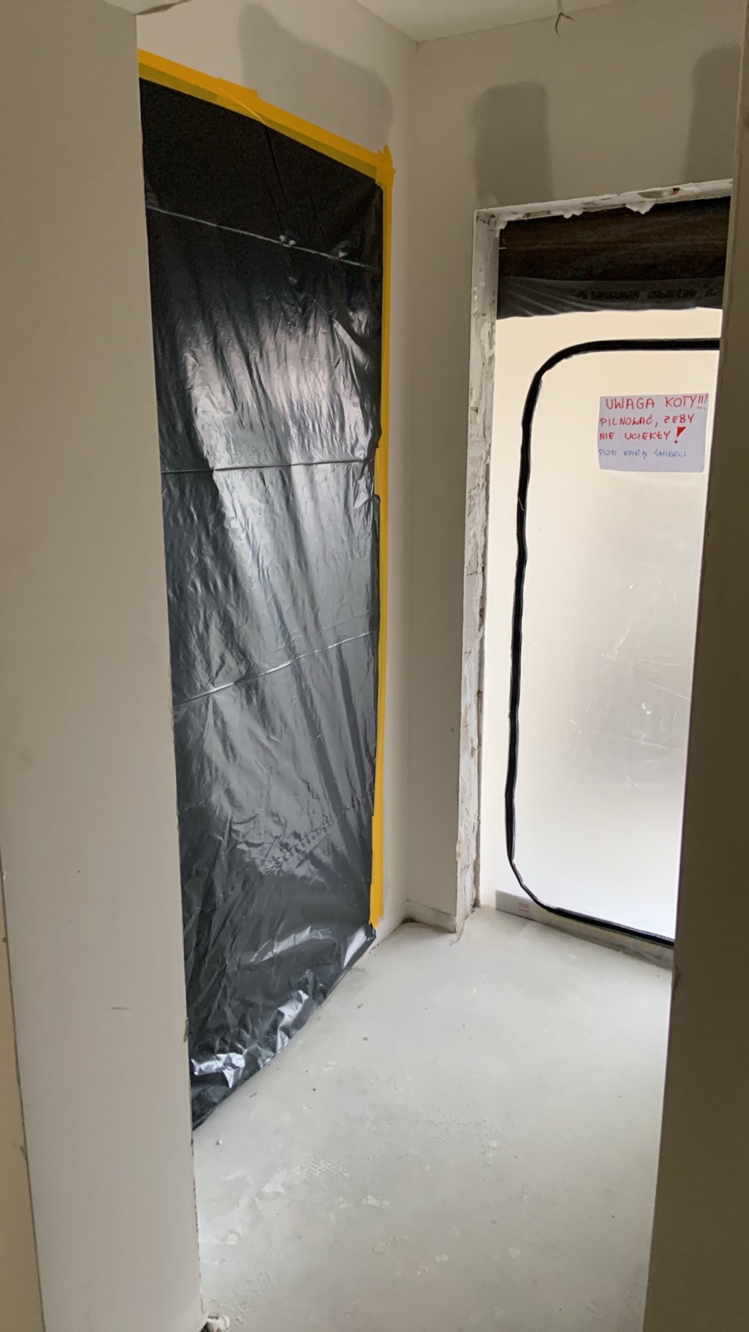 Przygotowanie do prac wykończeniowych: zabezpieczone folią i taśmą drzwi, świeżo wylana posadzka, kartka z ostrzeżeniem dla kotów. Wnętrze w trakcie prac.