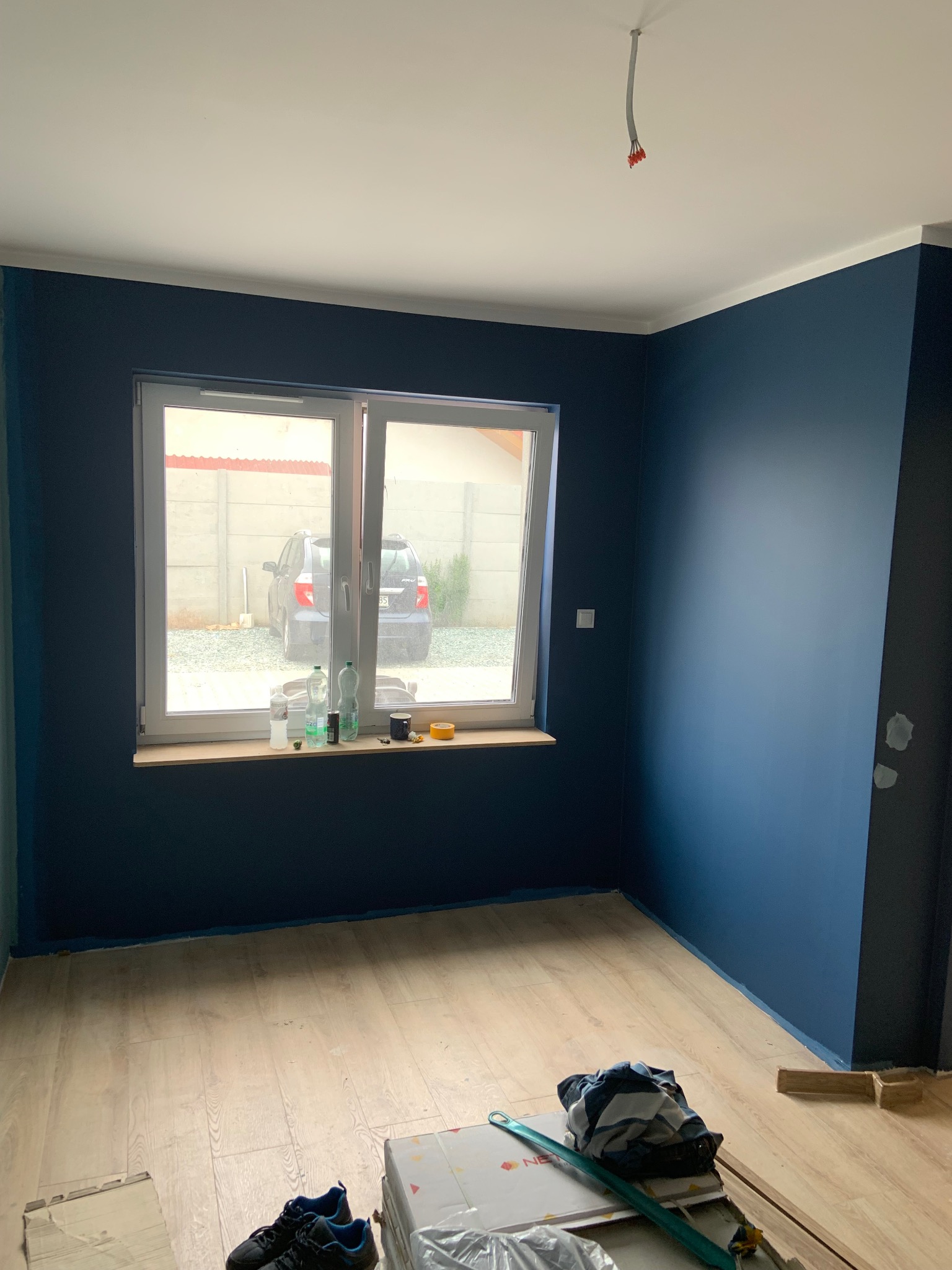 Pomieszczenie po malowaniu ścian na granatowo, widoczne narzędzia malarskie i podłoga z paneli. Przez okno widać samochód i betonowy płot.