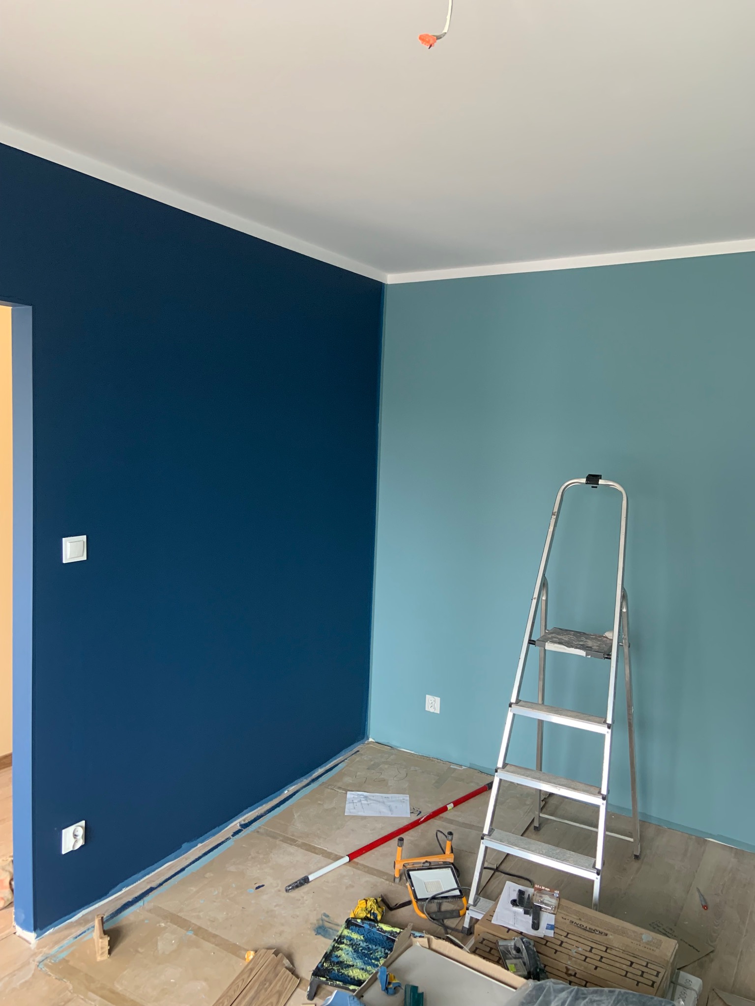 Ściana pomalowana na dwa kolory: granatowy i błękitny. Widoczna drabina malarska, wałek i inne narzędzia malarskie. Podłoga zabezpieczona folią malarską.