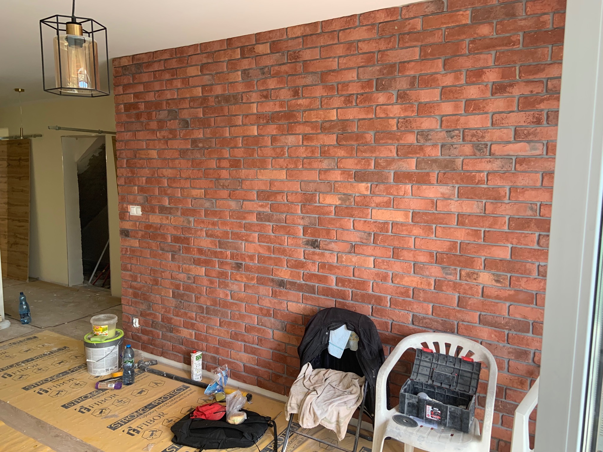 Ściana wyłożona czerwoną cegłą dekoracyjną, widoczna lampa wisząca i narzędzia pracy na podłodze zabezpieczonej kartonem. W tle widoczne otwarte drzwi.