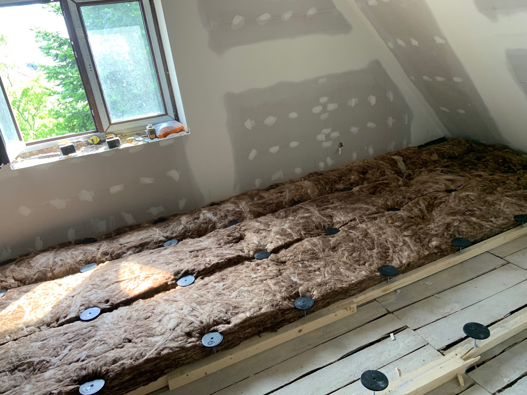 Ocieplenie poddasza wełną mineralną, widoczne mocowania. Ściany przygotowane do szpachlowania, okno dachowe z widokiem na zieleń. Prace wykończeniowe na poddaszu.