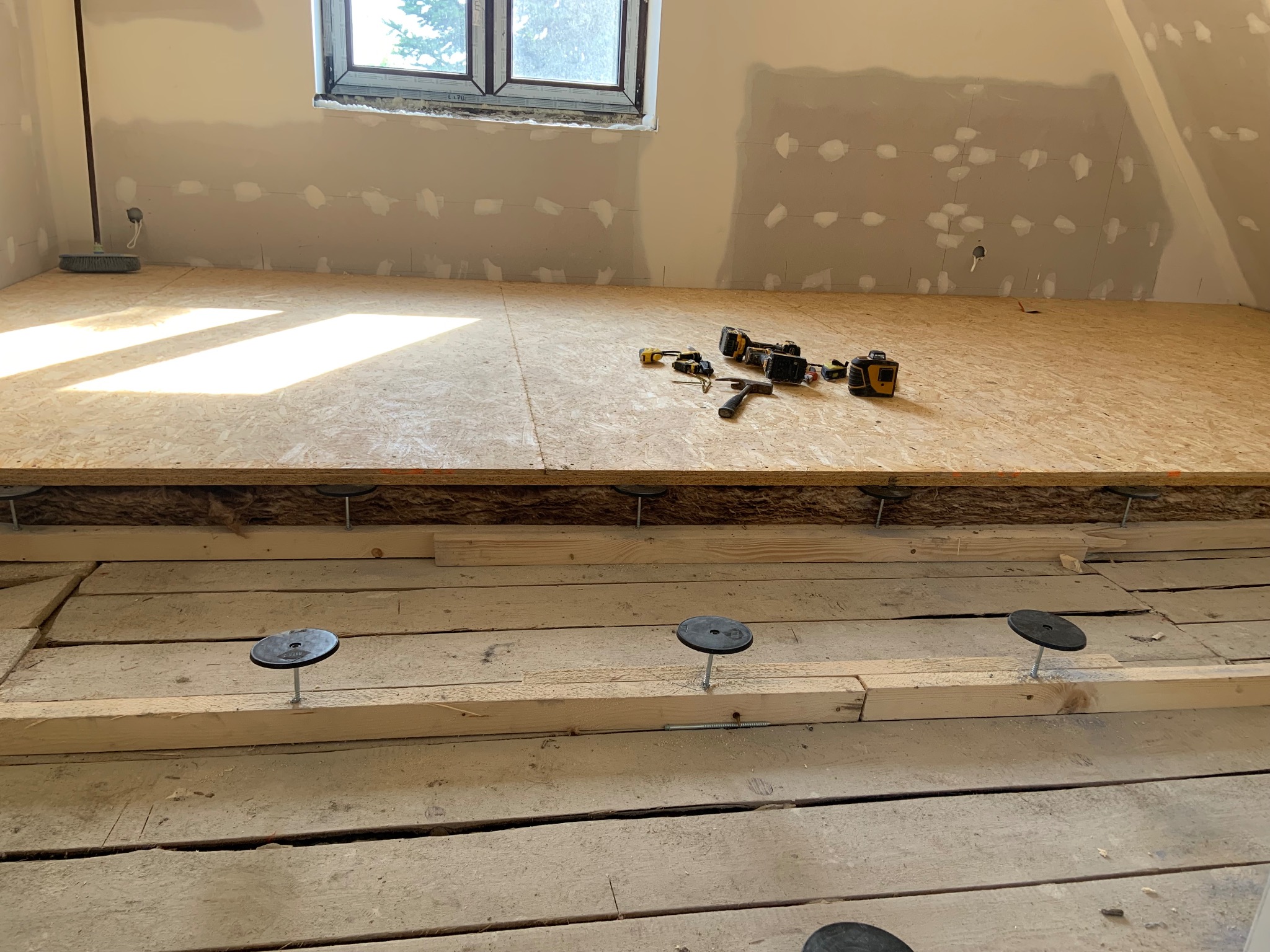 Poddasze w trakcie adaptacji: drewniana podłoga na legarach z regulowanymi wspornikami, narzędzia na płycie OSB, ściany przygotowane do szpachlowania.