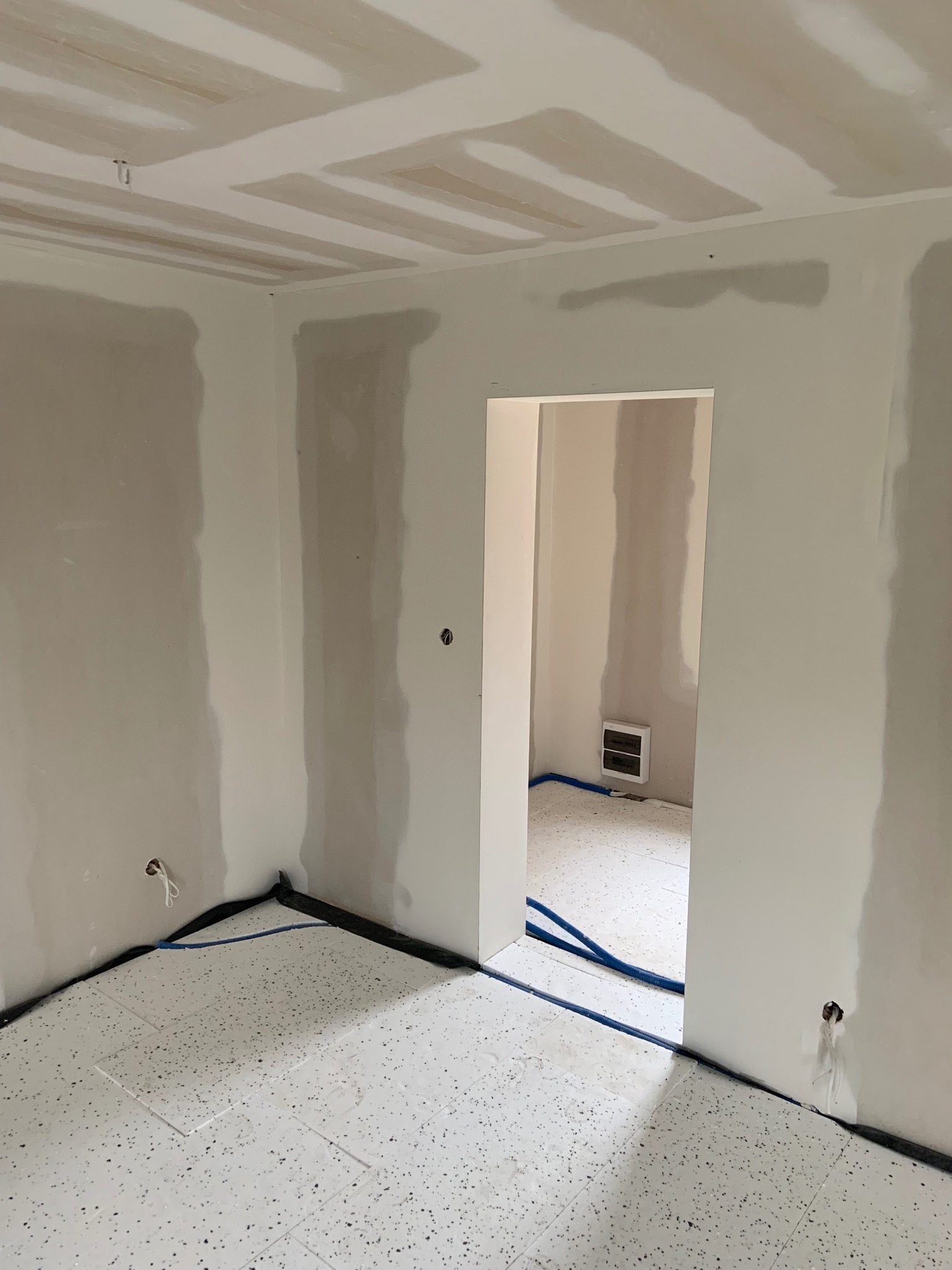 Wnętrze w trakcie prac wykończeniowych: ściany i sufit po szpachlowaniu, widoczne przewody elektryczne i rury. Na podłodze położona terakota.