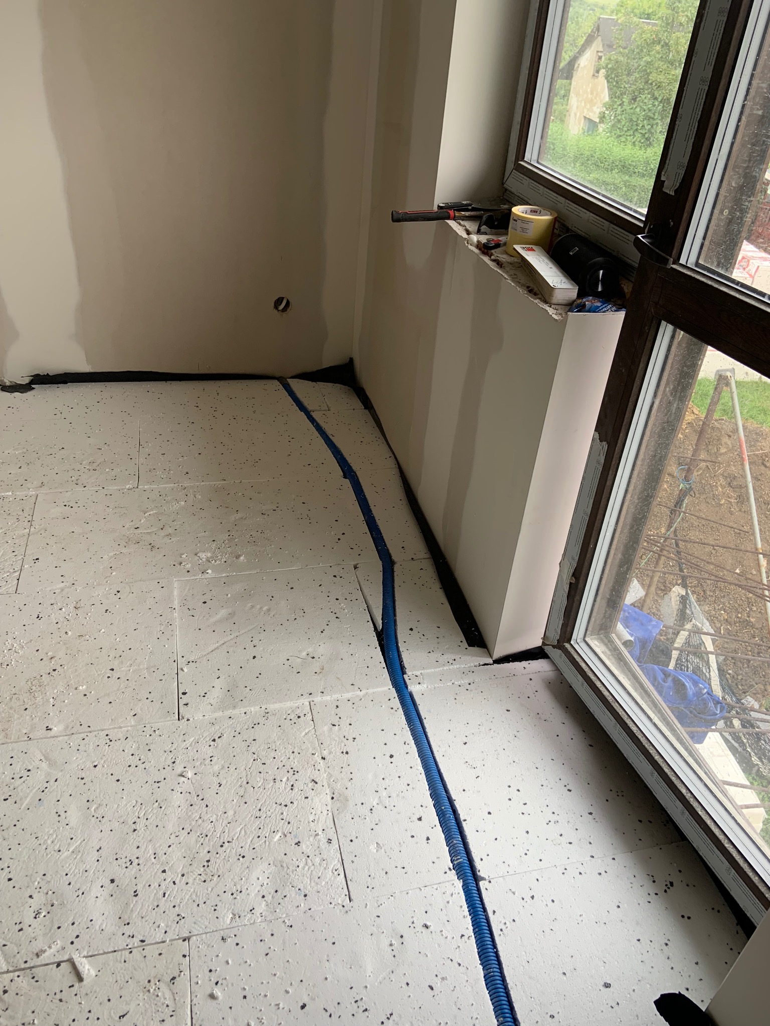 Przygotowanie podłoża: ułożone płyty izolacyjne na podłodze, niebieska rura instalacyjna, widok na okno z narzędziami na parapecie. Etap prac wykończeniowych.