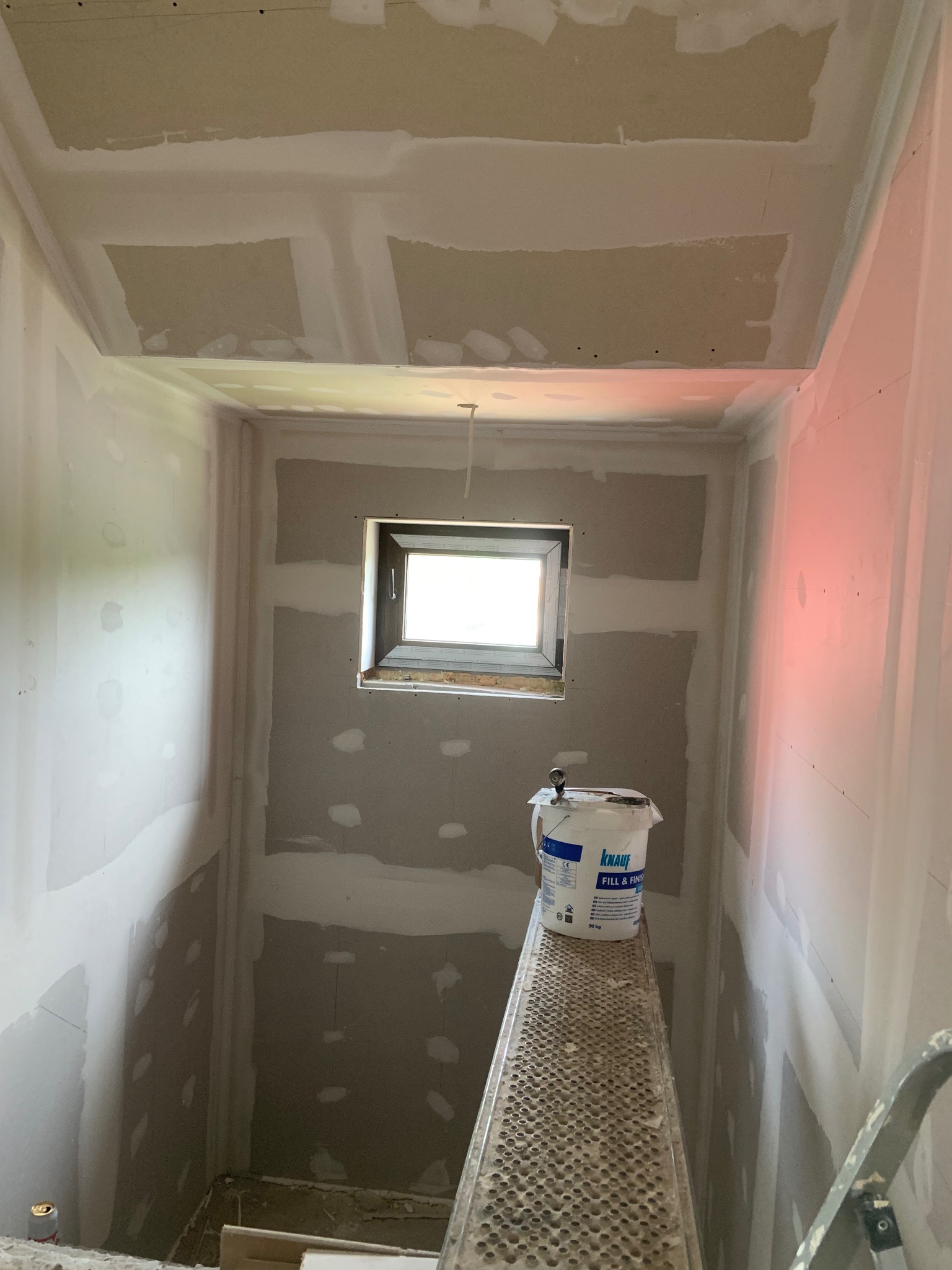 Wnętrze w trakcie prac przygotowawczych: ściany i sufit pokryte płytami gipsowo-kartonowymi, widoczne łączenia szpachlowane, wiadro z masą szpachlową na grzejniku.