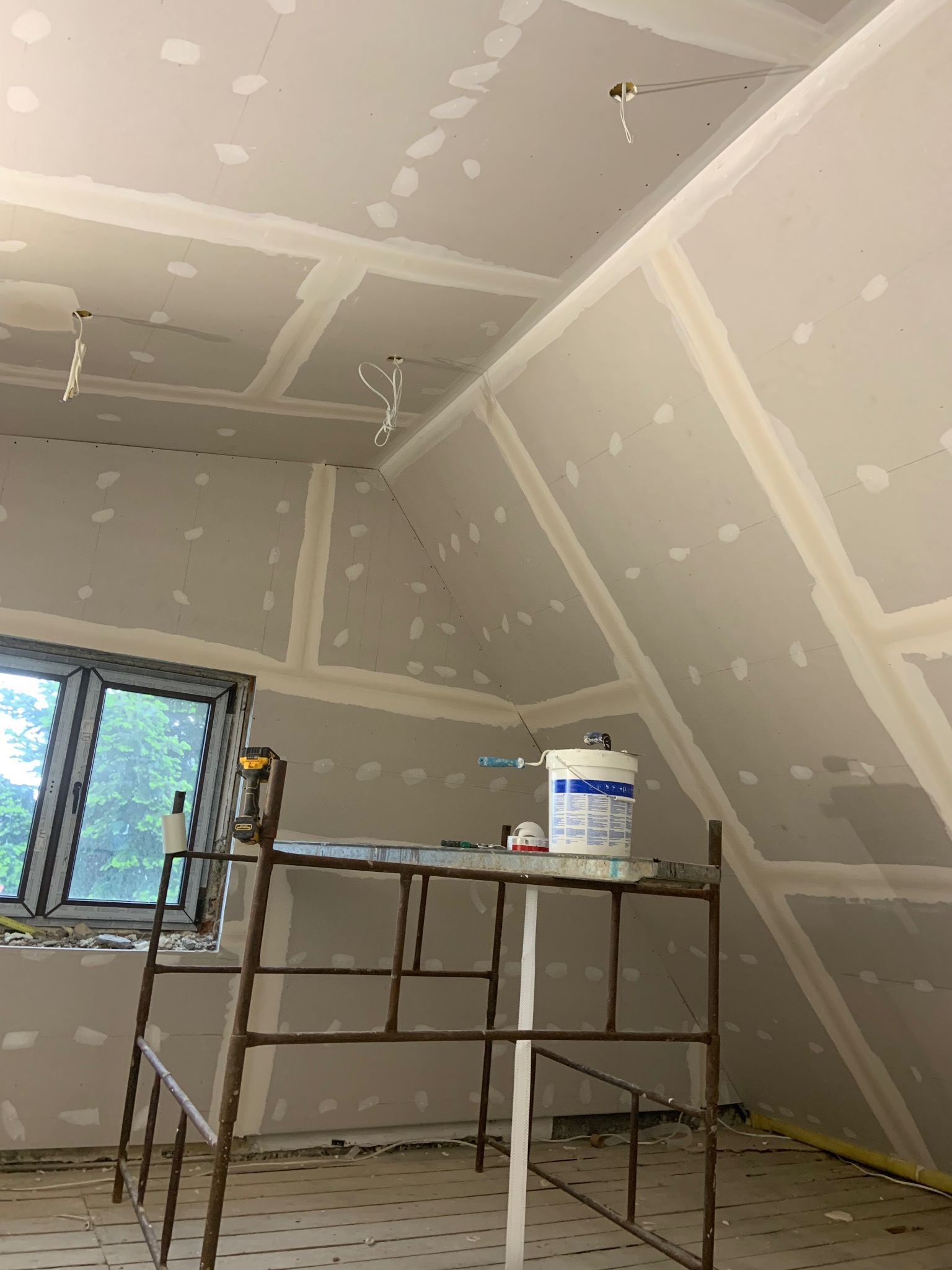 Poddasze w trakcie prac wykończeniowych: płyty gipsowe na skosach i suficie, szpachlowane łączenia, rusztowanie z narzędziami i farbą przy oknie. Widoczne kable oświetleniowe.