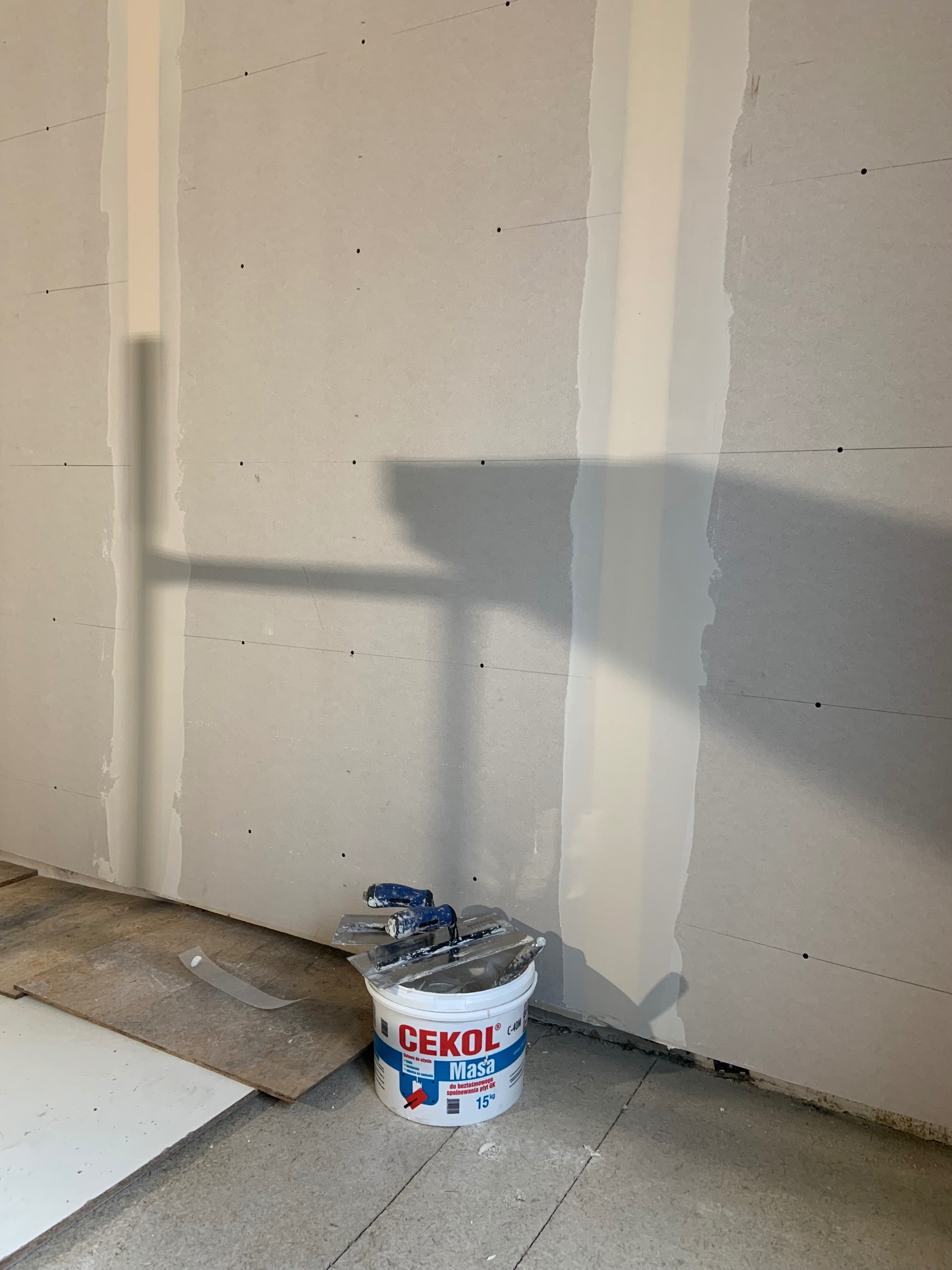 Ściana z płyt gipsowo-kartonowych, częściowo wyszpachlowana, z widocznym wiadrem z masą szpachlową i narzędziami. Etap prac wykończeniowych.