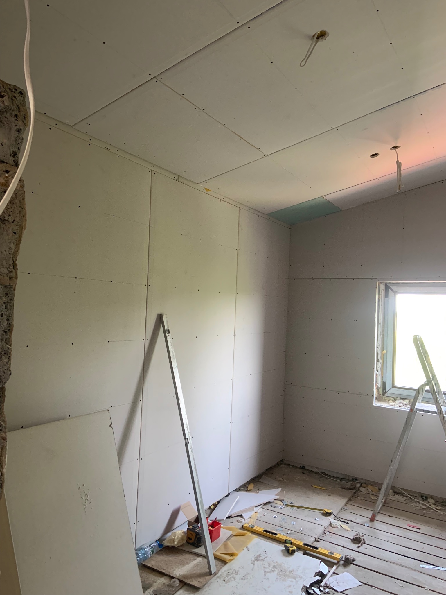 Pokój w trakcie montażu płyt gipsowo-kartonowych na ścianach i suficie. Widoczne narzędzia, drabina i fragment odsłoniętej ściany z cegły. Oświetlenie dzienne przez okno.