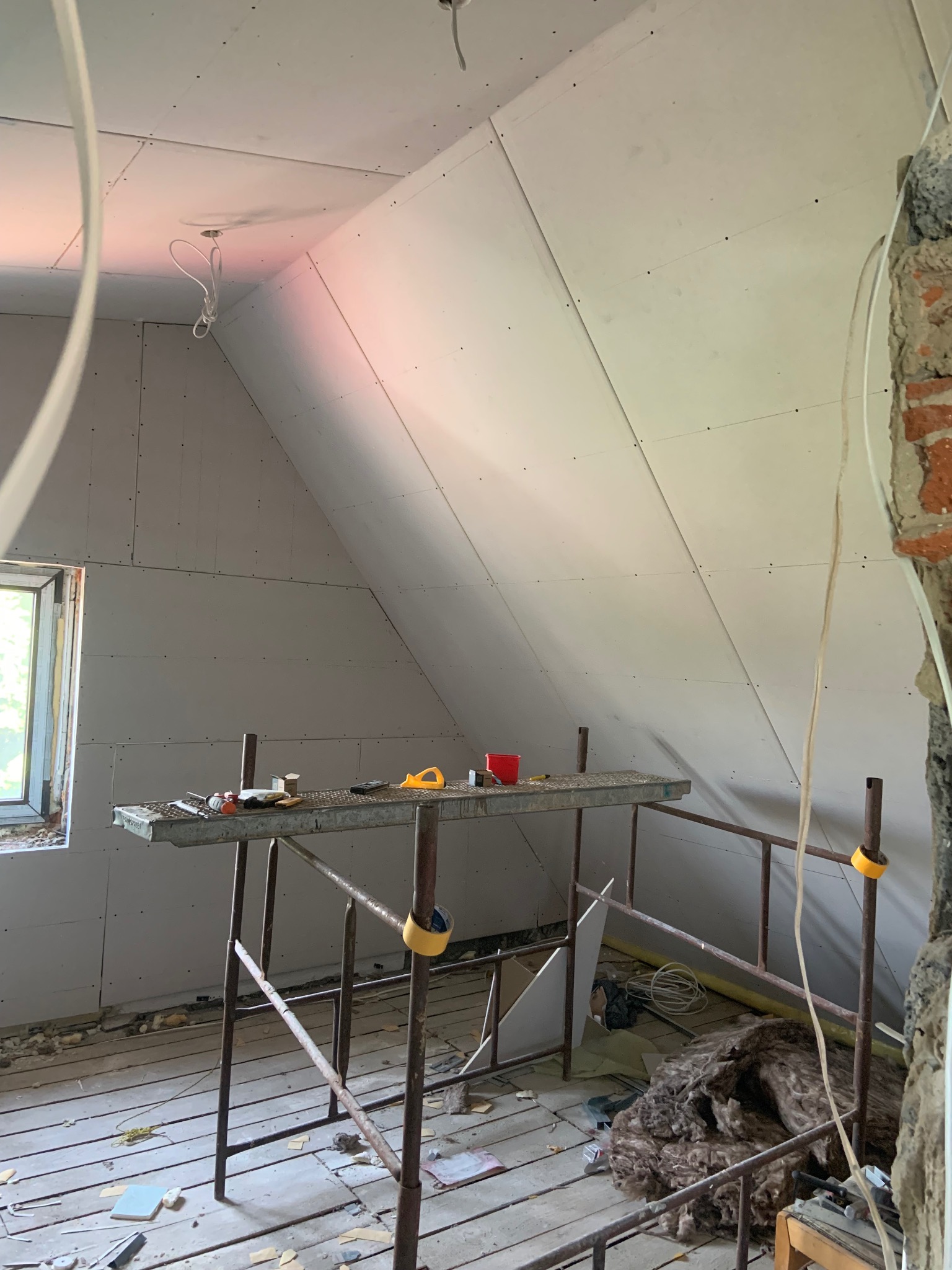 Poddasze w trakcie prac wykończeniowych: płyty gipsowe na ścianach, rusztowanie z narzędziami, widoczne kable i izolacja. Fragment ściany z cegły.