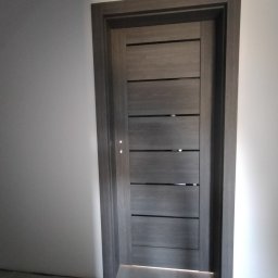 Tomasz koczorowski - Zamknięte, nowoczesne drzwi wewnętrzne w odcieniach szarości z poziomymi, ciemnymi akcentami, osadzone w szarej futrynie, na tle białej ściany i szarej podłogi.