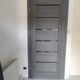 Tomasz koczorowski - Szare, nowoczesne drzwi wewnętrzne z poziomymi panelami, zamontowane w odnowionym wnętrzu. Widoczny fragment grzejnika i gniazdko elektryczne.