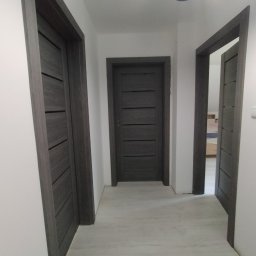 Tomasz koczorowski - Wnętrze korytarza z trzema ciemnoszarymi drzwiami z poziomymi panelami, białe ściany i jasna podłoga. Widoczna lampa sufitowa.