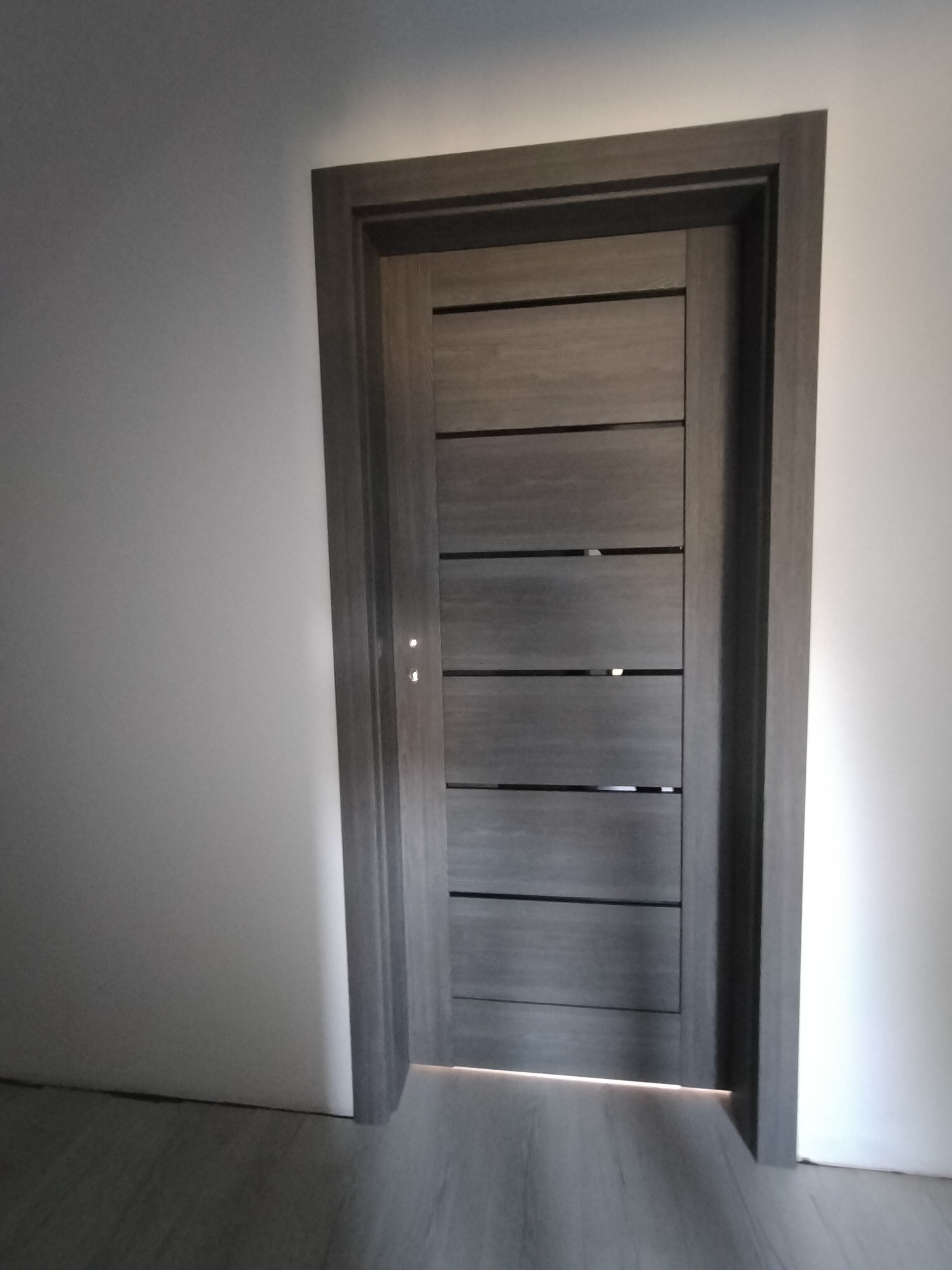 Zamknięte, nowoczesne drzwi wewnętrzne w odcieniach szarości z poziomymi, ciemnymi akcentami, osadzone w szarej futrynie, na tle białej ściany i szarej podłogi.