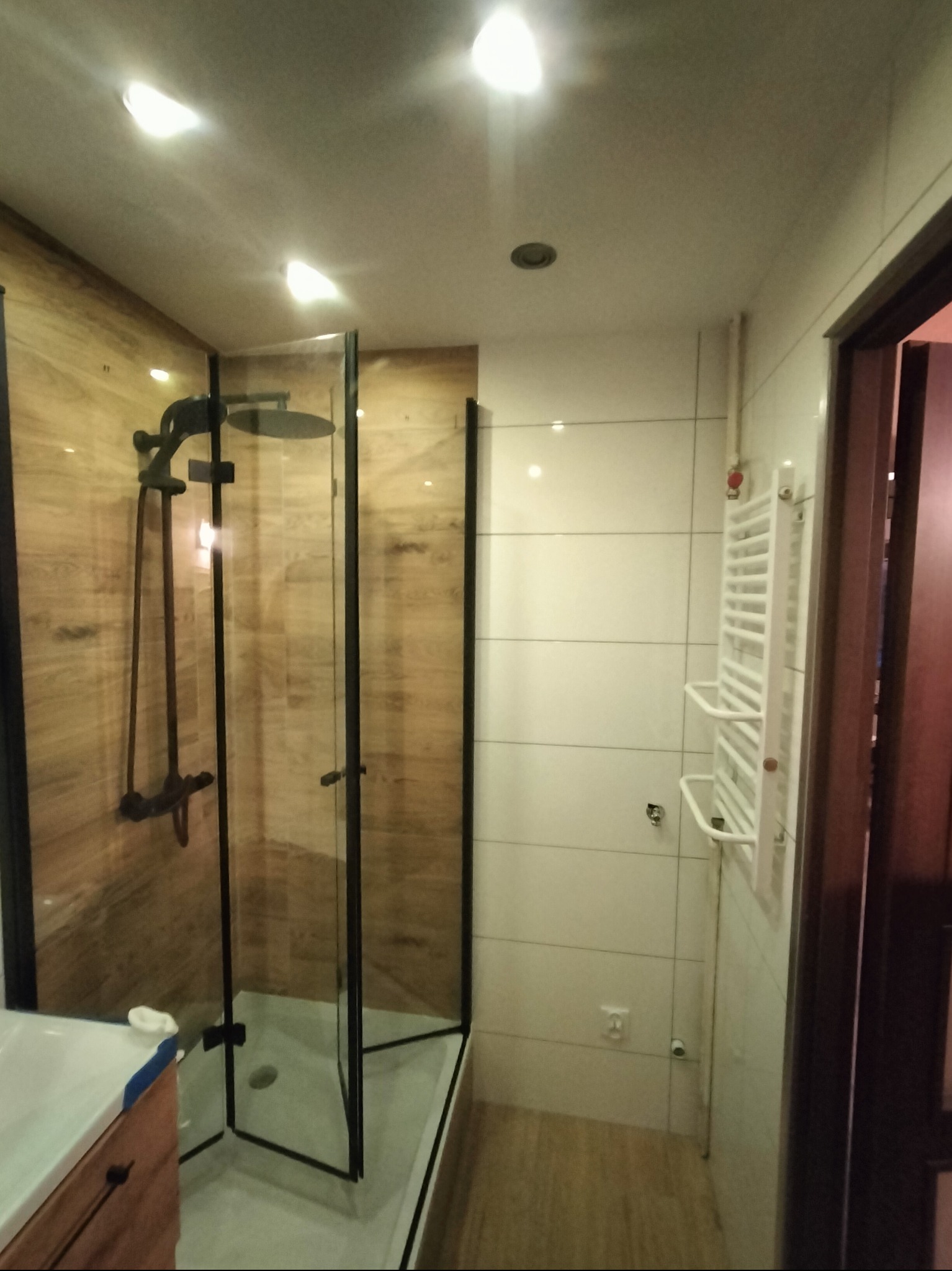 Nowoczesna łazienka z kabiną prysznicową typu walk-in z czarnymi profilami, drewnopodobnymi płytkami i białymi płytkami na ścianie z grzejnikiem drabinkowym.