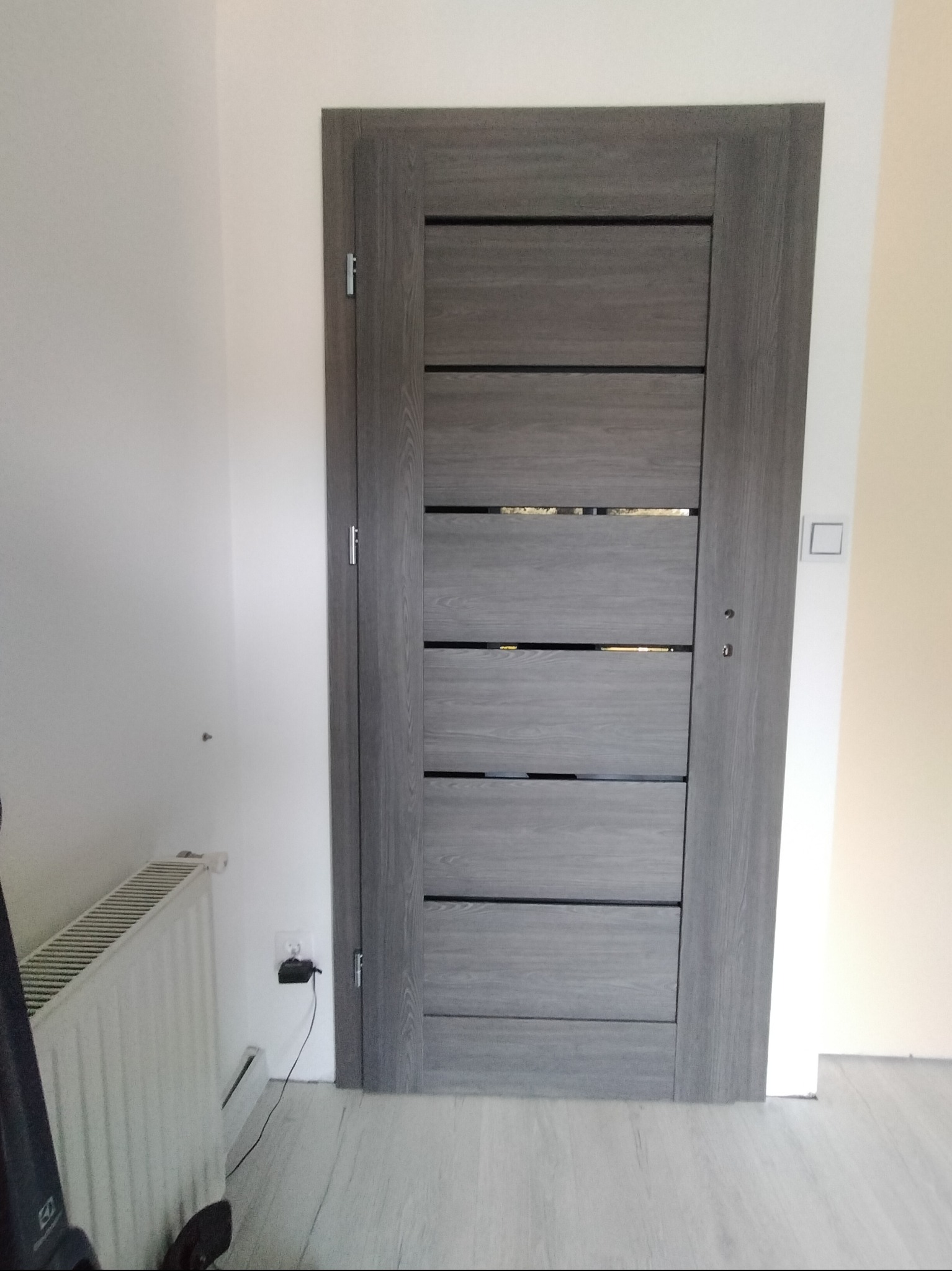 Szare, nowoczesne drzwi wewnętrzne z poziomymi panelami, zamontowane w odnowionym wnętrzu. Widoczny fragment grzejnika i gniazdko elektryczne.