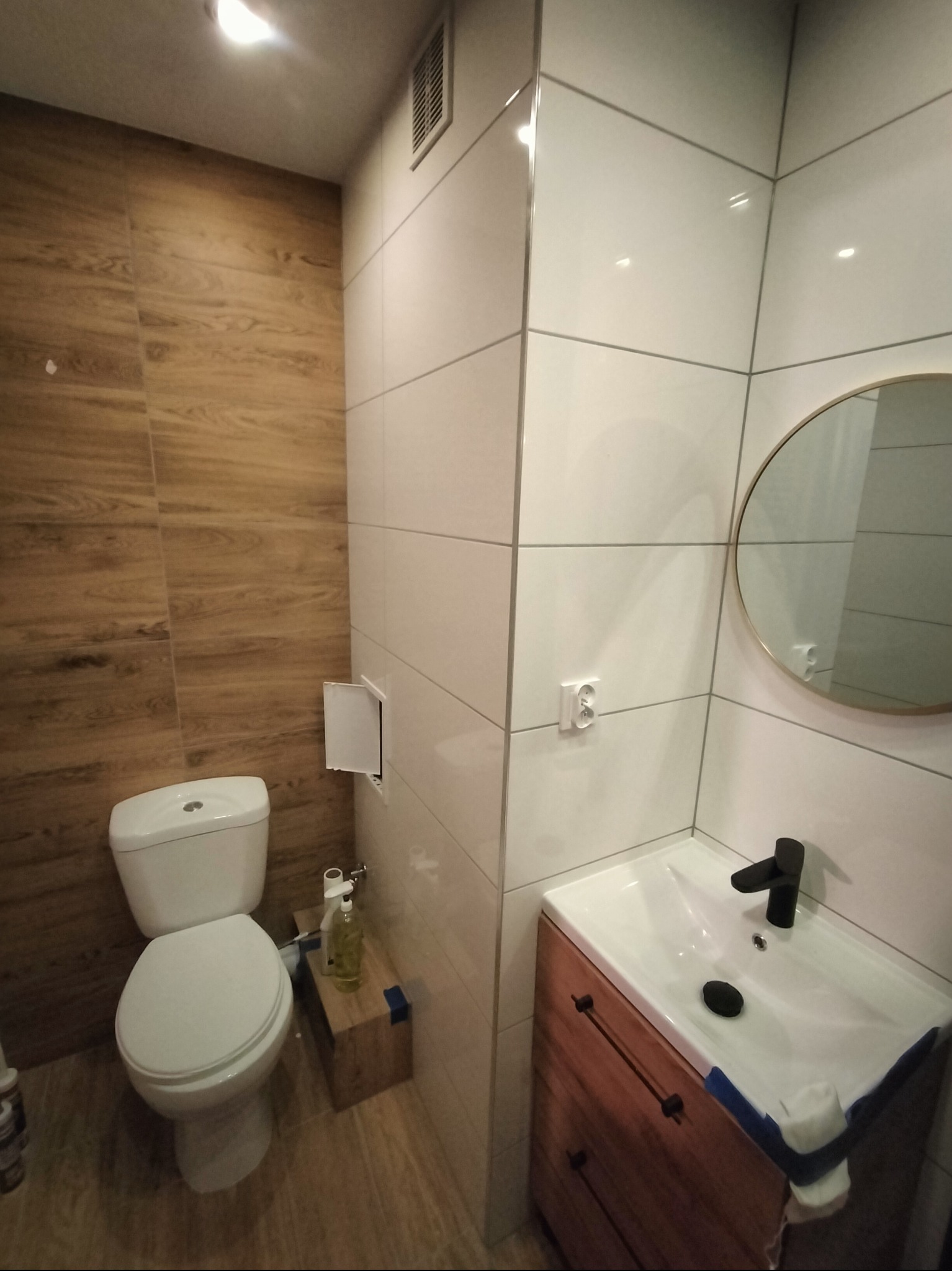 Nowoczesna łazienka po remoncie: białe i drewnopodobne płytki, okrągłe lustro, umywalka z szafką. Widoczne detale wykończenia i armatura.