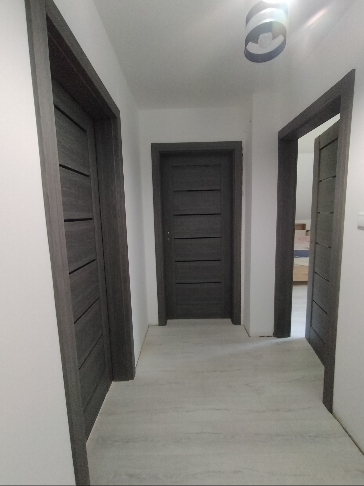 Wnętrze korytarza z trzema ciemnoszarymi drzwiami z poziomymi panelami, białe ściany i jasna podłoga. Widoczna lampa sufitowa.