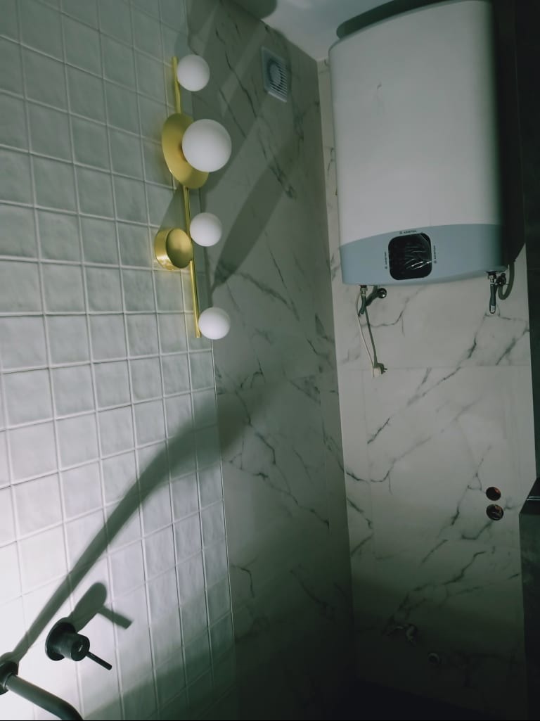 Wykończona łazienka z białymi płytkami, marmurową ścianą i złotą lampą. Widoczny bojler. Ujęcie z dołu, podkreślające wysokość pomieszczenia i nowoczesny design.