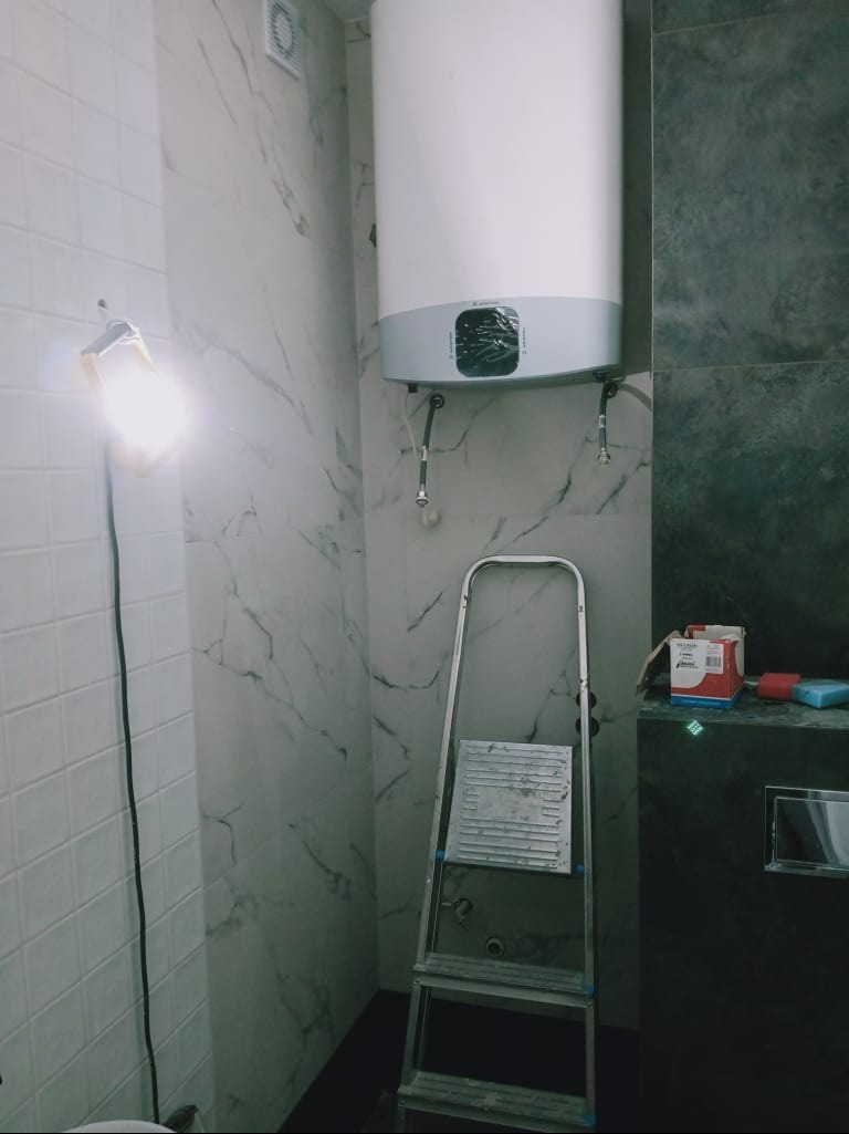 Montaż bojlera w łazience z marmurowymi płytkami. Widoczna drabina i lampa halogenowa. Etap prac wykończeniowych, z elementami instalacji wodnej.