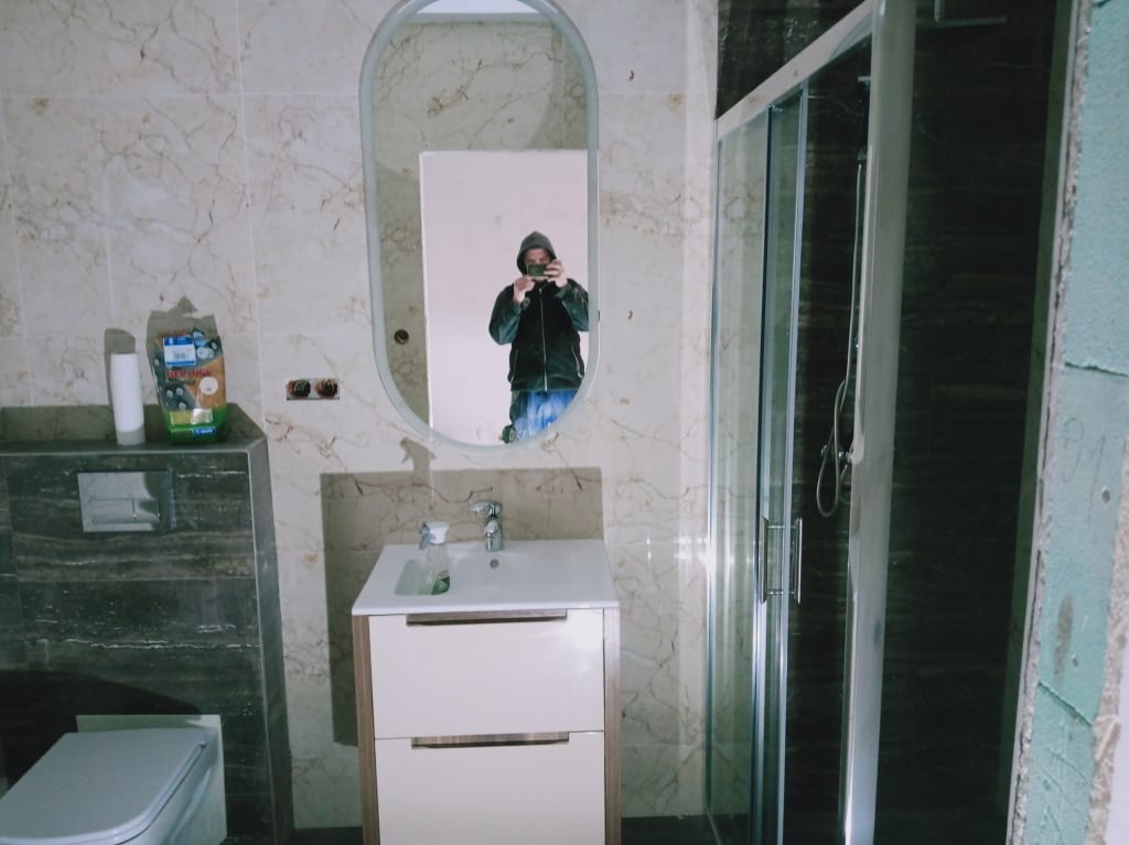 Wykończona łazienka z beżowymi płytkami imitującymi marmur, kabiną prysznicową i umywalką z szafką. W lustrze odbija się osoba robiąca zdjęcie.