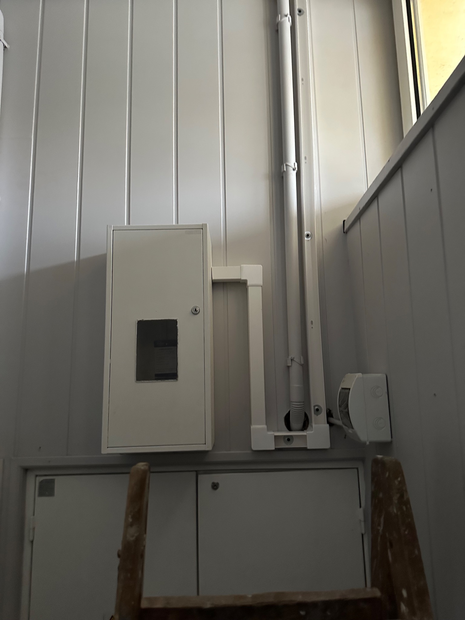 Biała skrzynka elektryczna z otworem, rura instalacyjna i puszka na ścianie z paneli. Fragment drabiny w dolnej części kadru. Ujęcie z dołu.